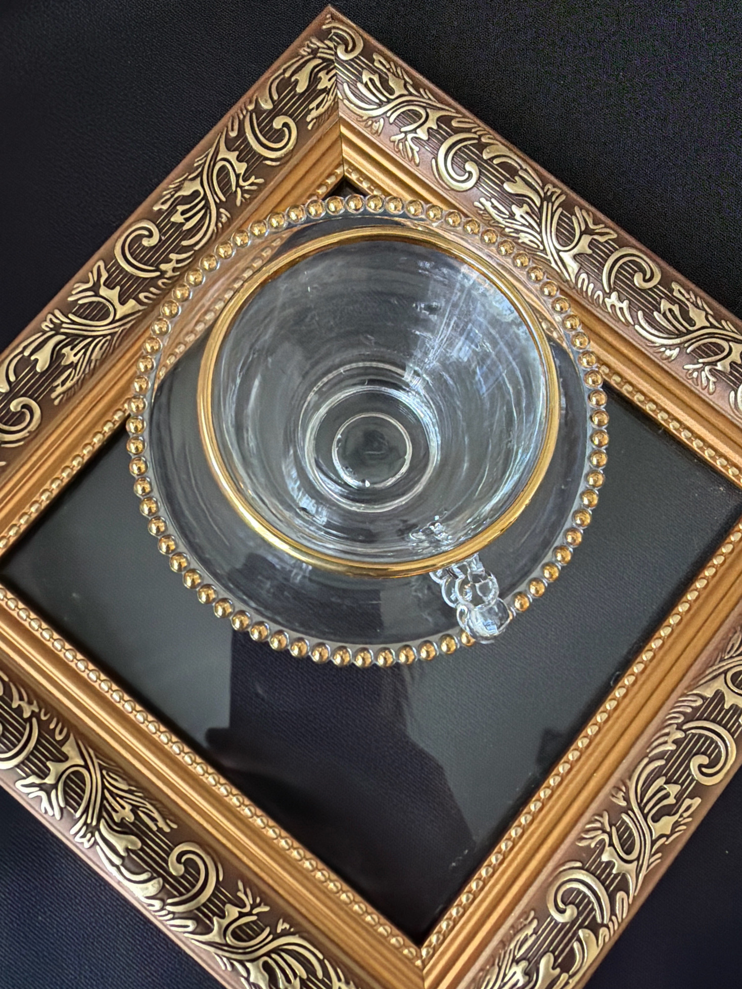 This is a close-up, high-angle shot of an ornate, golden picture frame tilted on a black surface. Inside the frame, centered, is a clear glass teacup and saucer, both with gold rims. The teacup is inverted, resting on its rim within the saucer. The saucer is also adorned with a beaded gold trim. The frame itself has a rich, dark brown base with intricate, raised gold floral and scrollwork patterns. The background is a textured black fabric, possibly felt or velvet. There are no people or animals visible, and no indication of a specific location in Бабініцкі сельскі Савет, Belarus. The scene is static and appears to be a still life arrangement.