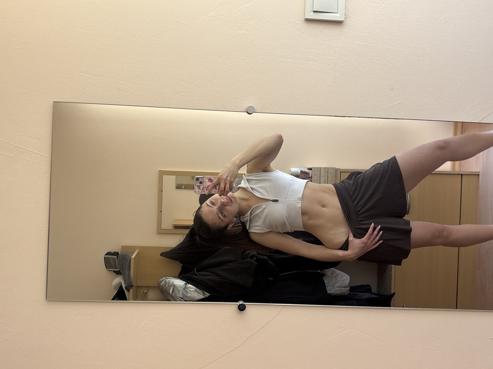A young woman is captured in a full-body mirror selfie, with the original image rotated 90 degrees counter-clockwise. She stands upright, facing the camera reflected in the mirror. She has long, dark hair and fair skin, looking directly into the smartphone she holds in her left hand. The phone has a floral or patterned case. She wears a white, short-sleeved crop top with a front zipper, exposing her midriff, and a dark brown or black pleated skirt or shorts. Her right hand rests gently on her hip. She has long, light-colored fingernails.
The large, rectangular mirror occupies most of the frame, reflecting a light beige or peach-colored textured wall. Above the mirror, a white light switch or outlet is visible on the wall. The wall continues below the mirror as well. Within the reflection, behind the woman, a wooden furniture piece, possibly a shelf or small cabinet, is visible. On it, a dark jacket is draped, alongside what appears to be a grey or silver puffy garment. Another smaller, framed mirror is reflected further back on the wall. The setting appears to be an indoor room, likely a private space such as a bedroom or dressing area, in Varna, Bulgaria. The lighting is consistent with indoor conditions, suggesting no specific time of day. No legible text is visible other than the abstract pattern on the phone case.