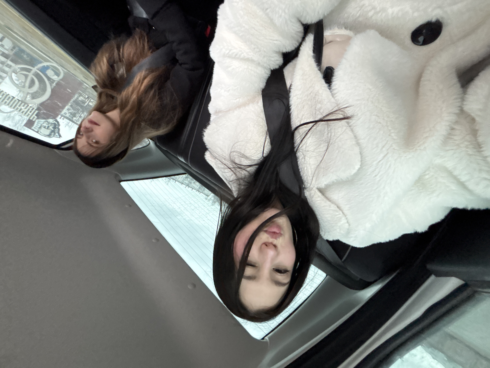 Two young women are inside a car. One woman is wearing a white, fluffy jacket and the other is wearing a dark coat. Both women are wearing seatbelts. The car appears to be parked and it looks to be a cloudy day outside. Based on the mirrored image reflected in the window, the location is likely the city of Chaikovsky, Russia.