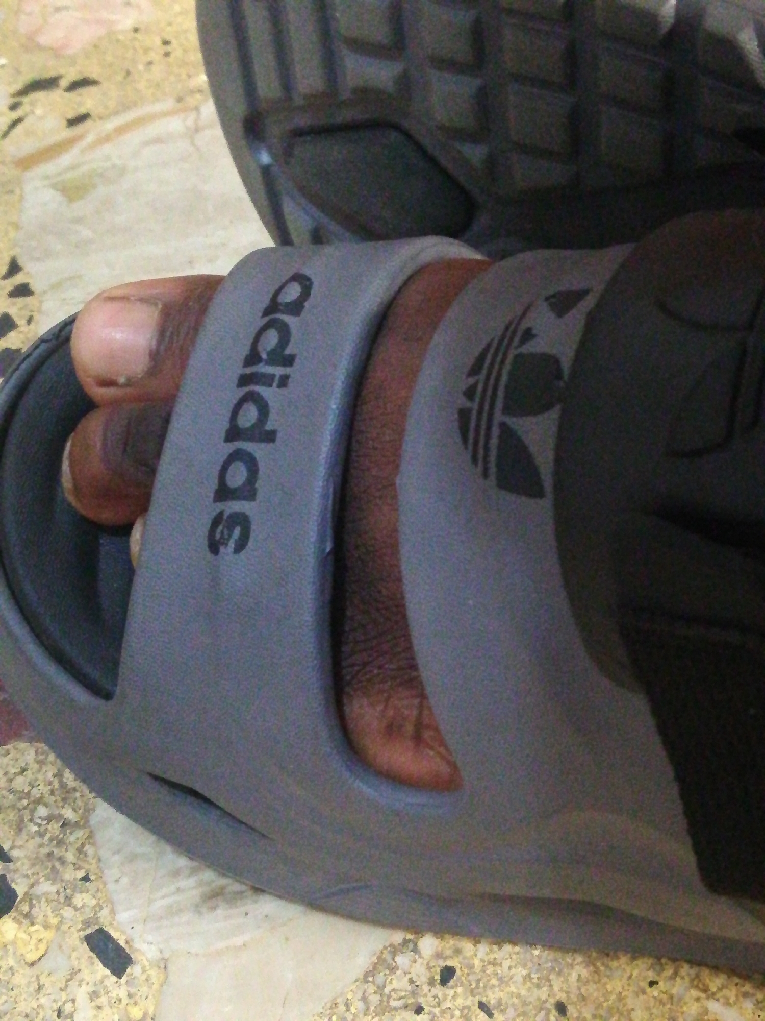 The image shows a close-up of a person's foot wearing a gray Adidas slide sandal. The sandal has the Adidas logo and the word "adidas" printed on it in black. The person's toes are visible. The background is a light-colored, patterned surface. No information indicates the location is in Lagos, Nigeria.