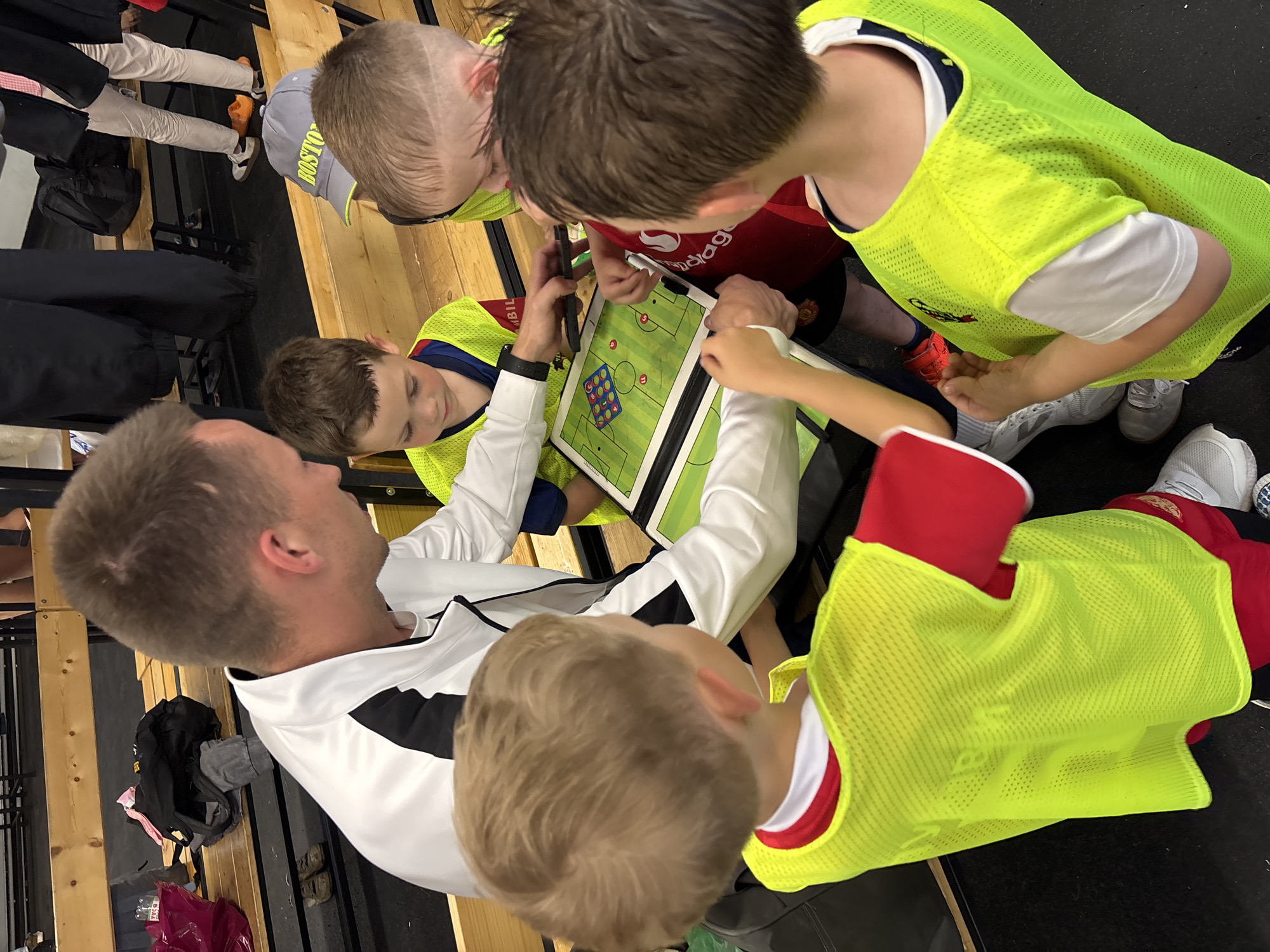 The image captures a group of individuals, likely a soccer coach and young players, gathered around a tactical board on a wooden bench in what appears to be a locker room or training facility. The coach, a man with short brown hair, is wearing a white and black jacket and is pointing to the board. He is surrounded by at least three young boys wearing bright yellow mesh vests over their team uniforms and red shirts. One boy is looking intently at the board, while another seems to be in conversation with the coach. The tactical board is green with white lines, depicting a soccer field with red and blue markers. In the background, other people are visible, including someone wearing light-colored pants and orange Crocs, and bags are placed on the benches. The environment suggests a casual, informal setting related to sports training. The location is not specified as Izhevsk, Russia, and no further information about the city or Russia is present in the image.