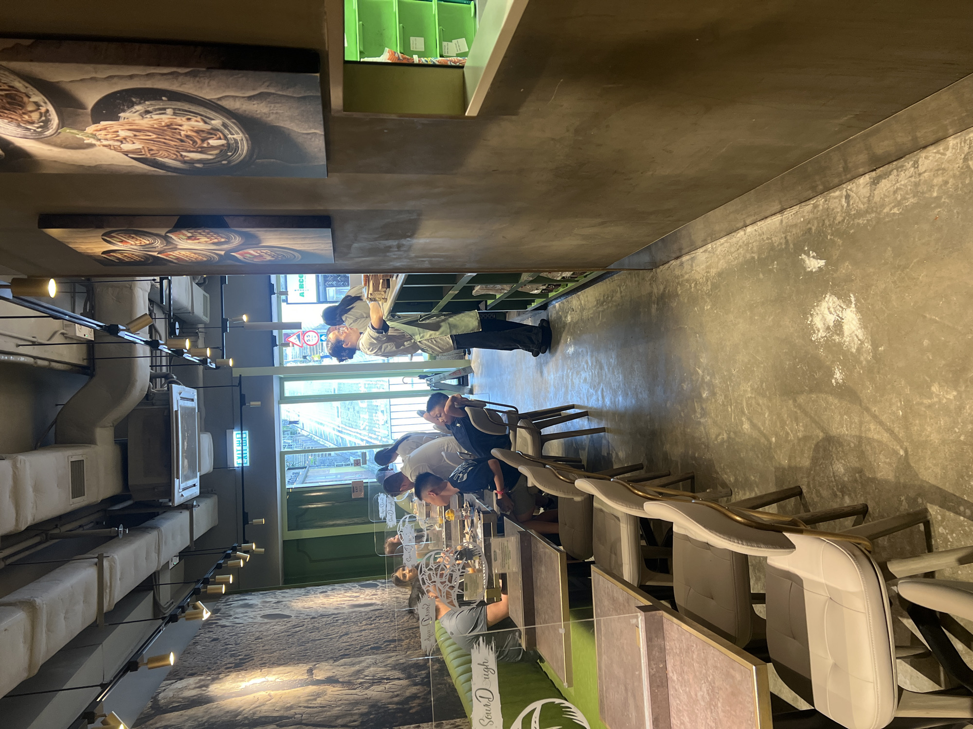 The image depicts the interior of a modern restaurant or cafe in Hong Kong, China. The scene captures several patrons seated at tables, engaged in dining or conversation. One person, a woman in a light-colored uniform with an apron, stands behind a counter or service area, appearing to be interacting with customers or preparing orders. The restaurant's decor features a textured wall with artwork depicting food, likely pasta dishes. There are also visible ceiling fixtures, air conditioning units, and lighting. Several tables are set with placemats and cutlery, and protective screens are in place between some seating areas. The flooring is a polished concrete. In the background, through large windows, a glimpse of an outdoor environment with trees and a street sign can be seen, suggesting an urban setting. An "EXIT" sign with Chinese characters is also visible. The overall atmosphere is casual and contemporary.