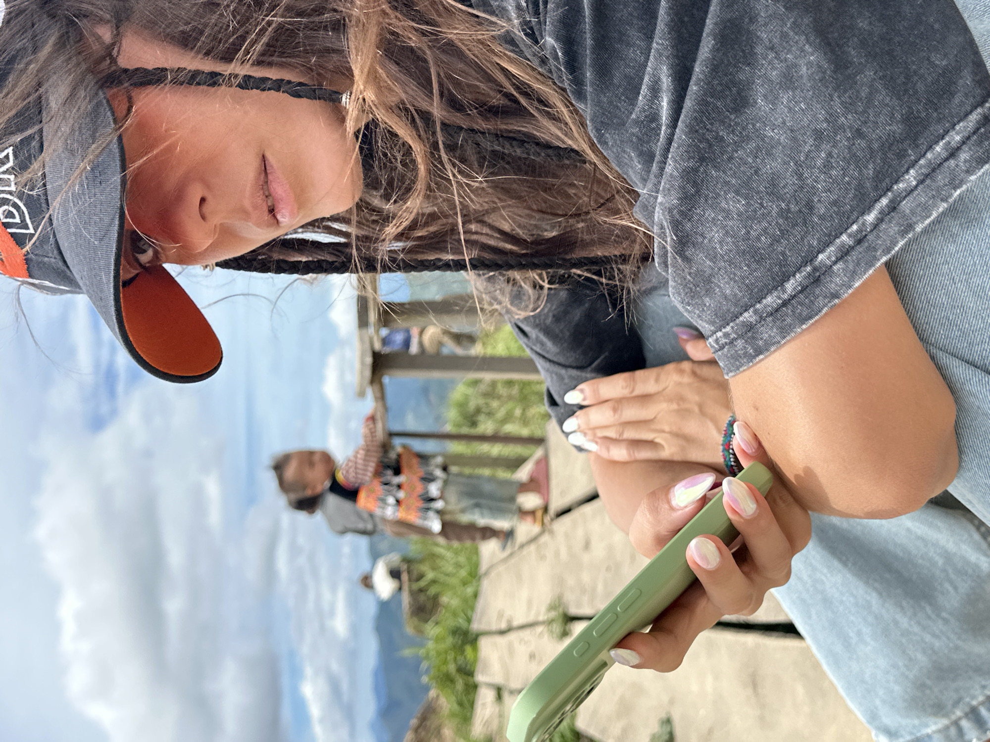 In the foreground, a woman wearing a dark grey, acid-washed t-shirt and light blue jeans is holding a light green smartphone in her left hand, which has iridescent, pearl-like fingernails. She also wears a dark baseball cap with an orange brim and a beaded bracelet on her right wrist. Her dark hair is styled in braids that hang down her shoulders and back. In the background, on a wooden walkway, a small child wearing a colorful traditional vest and light blue pants is standing and looking towards the left. The child is being supervised by an adult whose back is to the camera. The scene appears to be an outdoor viewpoint in Ta Xua, Vietnam, with lush green mountains and a cloudy sky visible in the distance.