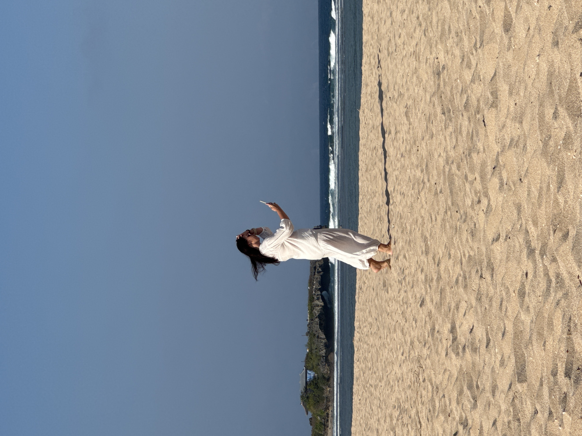 A lone person wearing loose white clothing walks on a sandy beach with the ocean in the background. The person is holding a white object, possibly a phone, up to their face. The ocean has moderate waves and a small island with a building on it is visible in the distance. The sky is clear and blue. The location is the beach in Kuta Selatan, Indonesia.