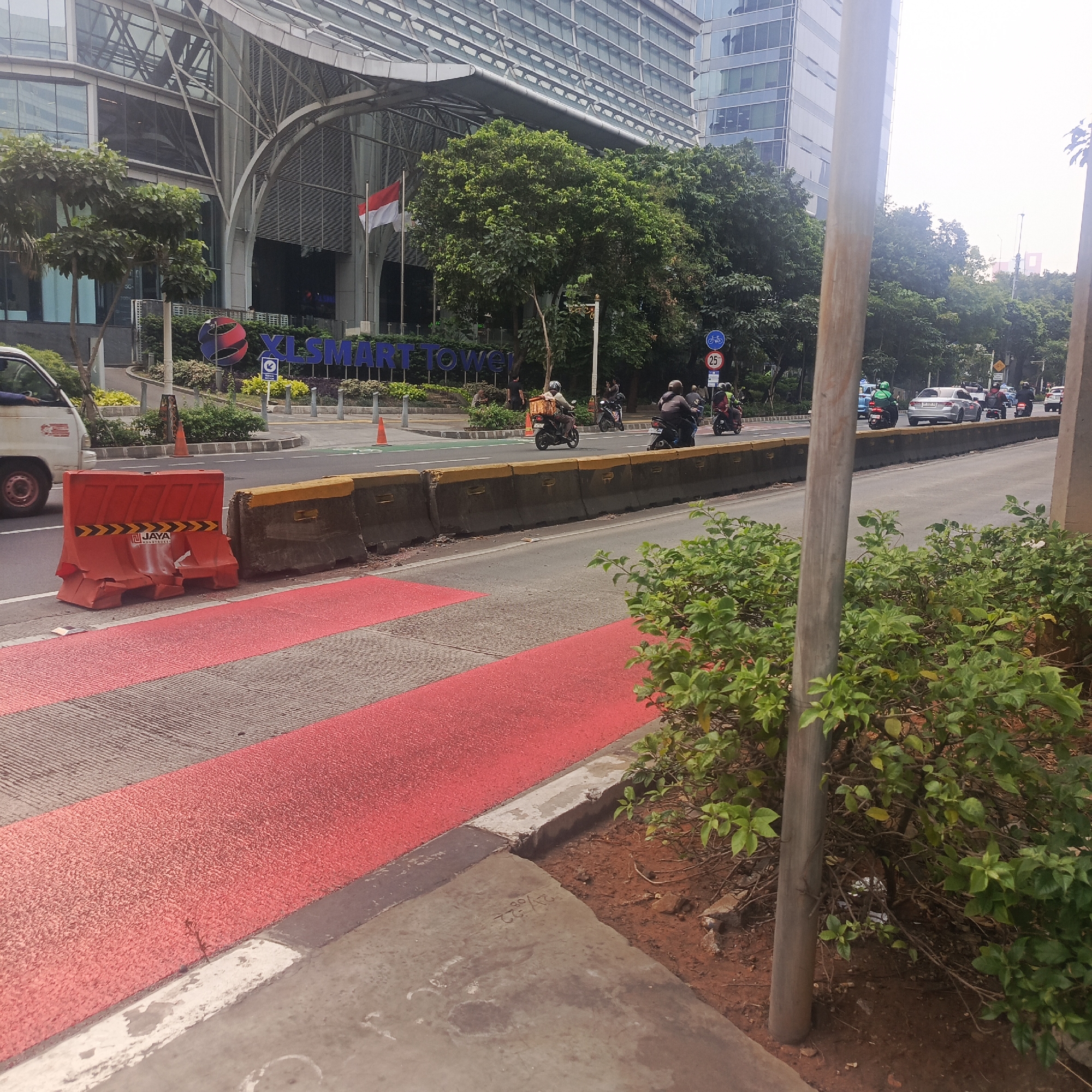 The image captures a street scene in South Jakarta, Indonesia, in front of the XL SMART Tower. The foreground features a pedestrian crossing with distinct red and grey markings. Orange plastic barriers and concrete barriers line the edge of the road, indicating potential construction or lane division. Several motorcycles, some with riders wearing helmets and one with a delivery basket, are present on the road, suggesting ongoing traffic. In the background, the modern architecture of the XL SMART Tower is visible, along with lush green trees and the Indonesian flag flying. The sky is overcast, creating a diffused light. The scene depicts a busy urban environment with a mix of infrastructure, vehicles, and greenery.