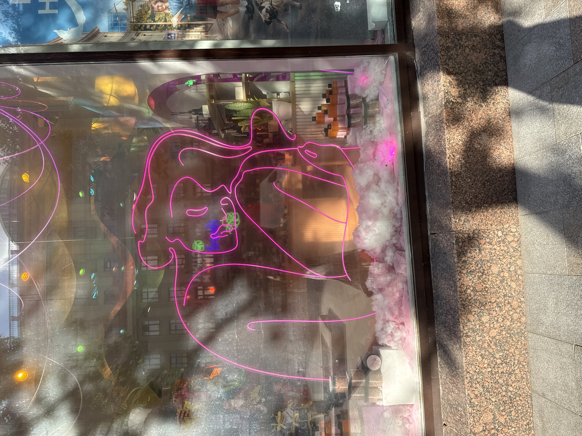 The image shows a storefront window display featuring a large, abstract neon pink outline of a figure with arms raised. The figure's outline is abstract and stylized, with a rounded head and two prominent arms extending upwards. Inside the window, there are various retail items displayed on shelves and pedestals. In the foreground of the display, there are stacks of clear jars filled with a light-colored granular substance, and several orange-bottled products. The display is enhanced with fluffy white and pink material, possibly cotton or fabric, arranged to create a soft, cloud-like effect, illuminated by pink neon lights. Reflections of buildings, trees, and sky are visible on the glass of the storefront, indicating it is daytime and possibly a sunny day. On the right side of the window, a poster is visible, and a section of the pavement and a granite-clad building base are shown below the window, suggesting an urban setting. The overall impression is a visually striking and artistic retail display.