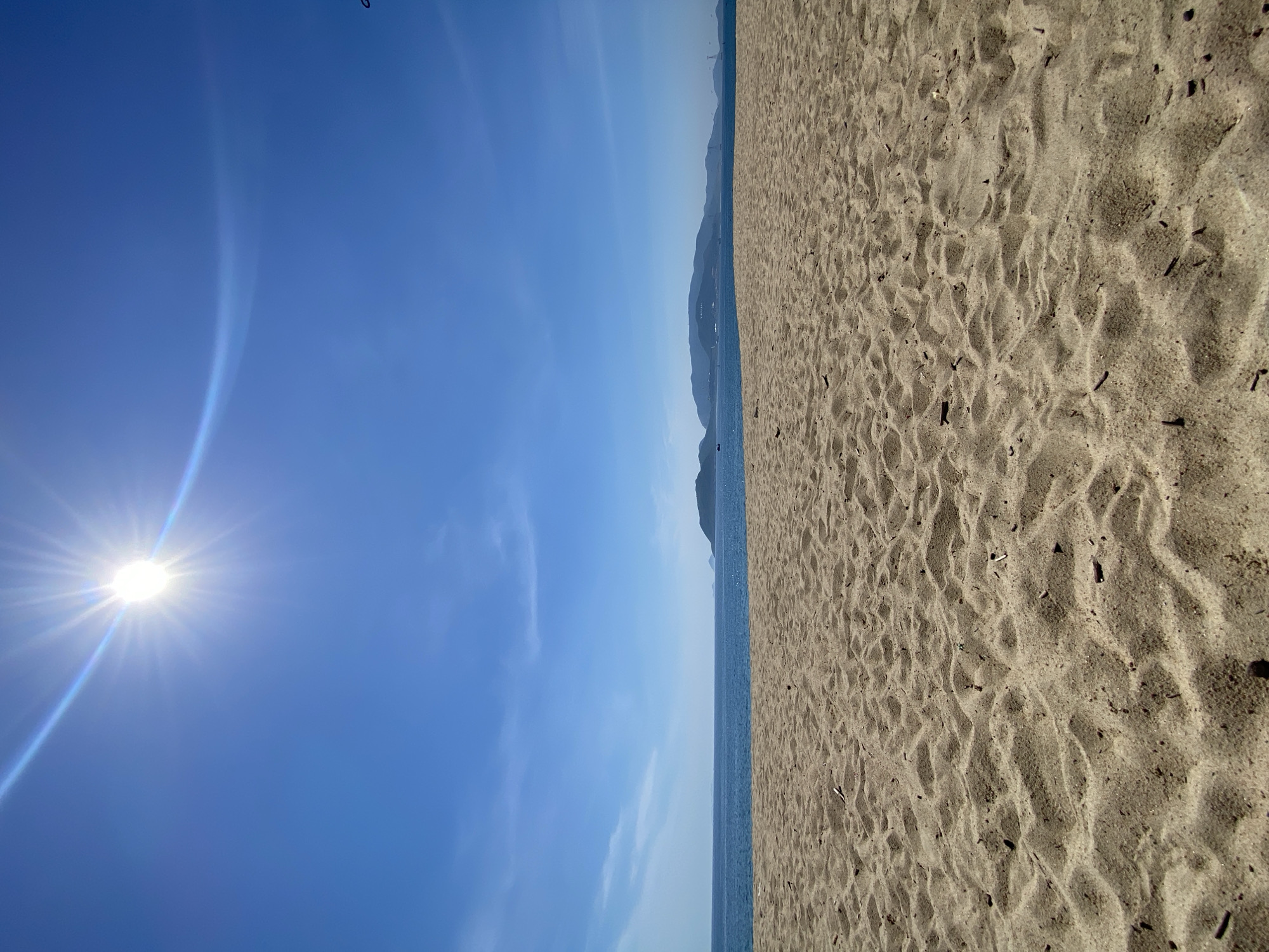 A low-angle shot captures a bright, sunny day at a beach. The foreground is dominated by rippled sand, with small pieces of debris scattered across it. In the mid-ground, the calm, blue ocean stretches to the horizon. Distant, hazy mountains form the backdrop, under a clear, vibrant blue sky. The sun is high in the sky, emitting a strong glare with visible rays. The overall scene suggests a tranquil beach environment in Nha Trang, Vietnam.