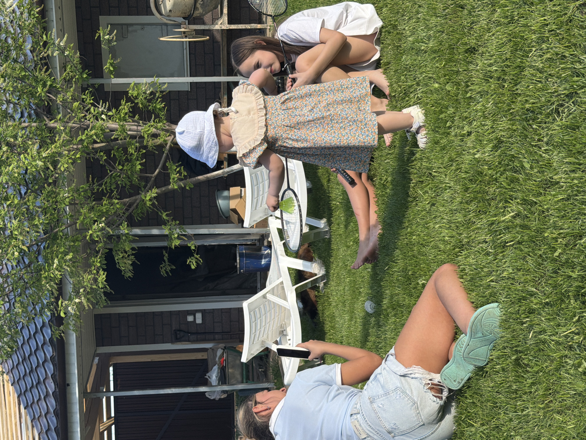 The image depicts a backyard scene in Chaikovsky, Russia. A woman lies on the grass, facing away from the viewer, looking at a smartphone. She is wearing a light blue short-sleeved shirt and ripped denim shorts. Her legs are extended, and she has on mint-colored sneakers. In the background, two girls are interacting. One girl in a white T-shirt is crouching down, looking at a younger girl in a blue floral dress and white sun hat. They both are holding badminton rackets. The background includes a residential building, white patio furniture, and a tree with white flowers. The lawn is green and well-maintained.