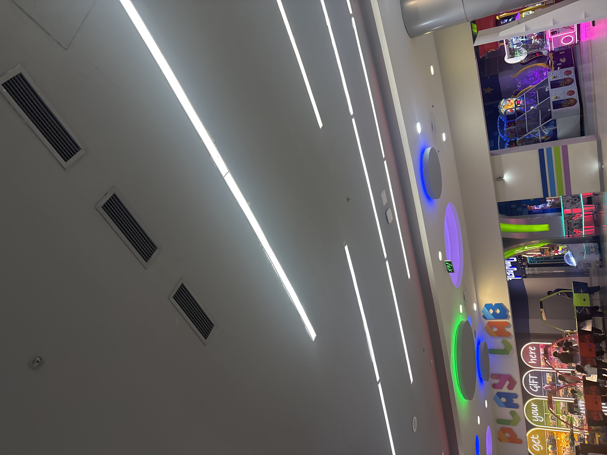 This image captures a vibrant indoor entertainment center, likely an arcade or family fun area, located in Yerevan, Armenia. The perspective is angled upwards, revealing a modern ceiling equipped with numerous long, linear LED light strips that provide bright, white illumination. Rectangular air vents and a circular smoke detector are also visible on the light-colored ceiling.
A prominent feature on the right side of the image is a large, brightly lit sign on a wall that spells out "PLAYLAB" in colorful, three-dimensional letters, incorporating shades of blue, green, orange, and purple. Directly below the main sign, smaller illuminated panels are arranged vertically, displaying the words "get," "your," "GIFT," and "here," indicating a prize or redemption area. The wall is further enhanced by large, circular, illuminated fixtures in blue, purple, and green, contributing to the lively atmosphere.
Beyond this main wall, a bustling arcade area is visible. It features various brightly lit arcade games, including basketball shooting games with orange hoops and what appear to be claw machines. Several individuals, comprising children and young adults, are actively engaged with the games, suggesting a fun and energetic environment. The overall setting is modern, designed for recreation, and filled with artificial lighting, making the specific time of day indistinguishable.