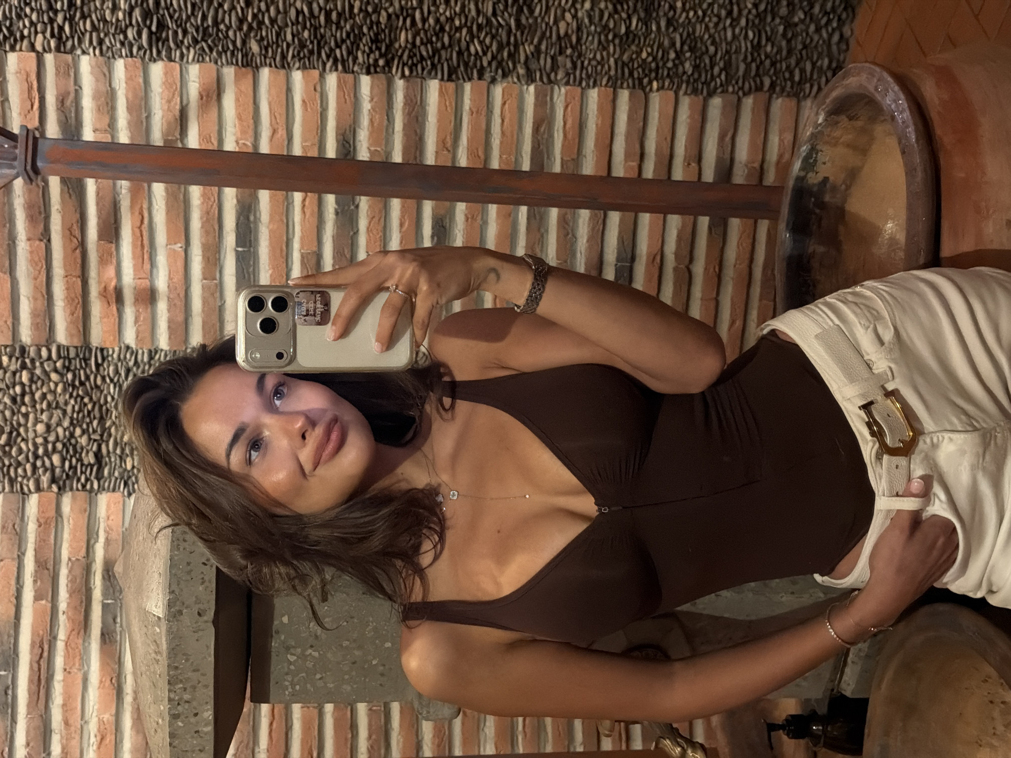 A woman is taking a selfie in what appears to be an interior setting with a decorative brick and stone wall. The background suggests a stylized rustic design. The woman is wearing a brown bodysuit and white pants. She is holding her phone in her left hand, taking the picture. The setting appears to be stylized but not identifiable enough to place specifically in Krasnoyarsk. There are large pots and what appears to be stone or concrete elements in the immediate surroundings.