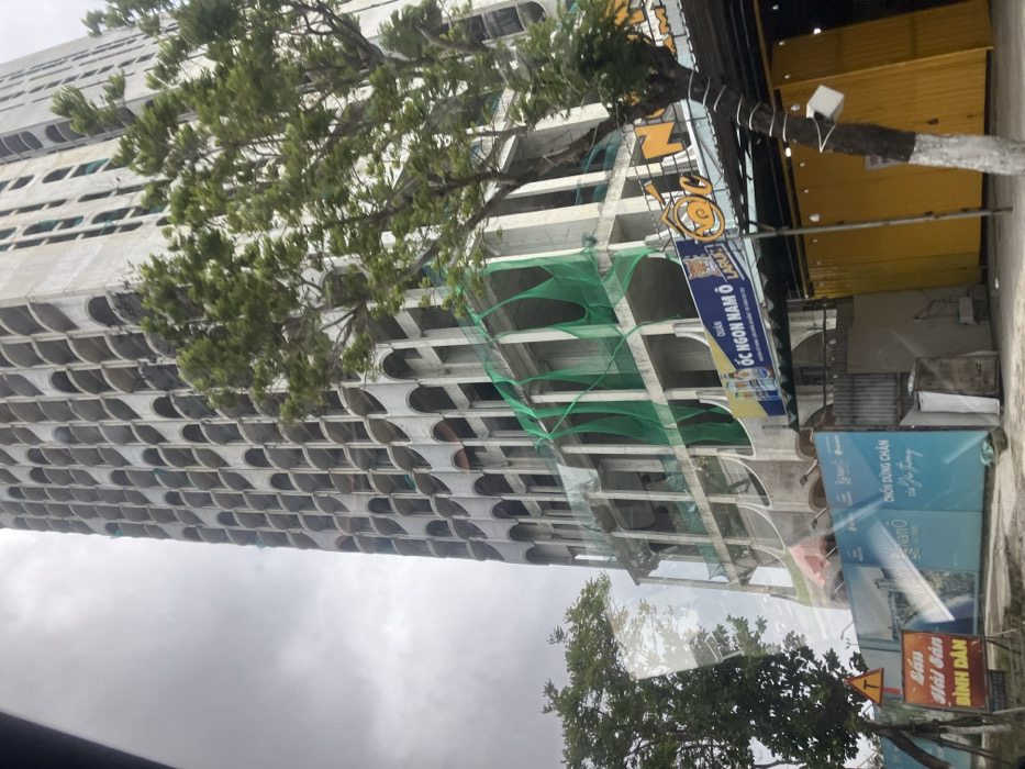 The image depicts a tall, multi-story concrete building under construction, viewed from a street-level perspective in Hải Vân Ward, Vietnam. The building's facade features a repeating pattern of numerous arched openings, suggesting balconies or window frames. Green safety netting is visible draped over several lower and mid-level sections of the unfinished concrete structure, particularly around exposed floor plates and columns.
In the foreground, a blue construction hoarding runs horizontally, displaying Vietnamese text including "Nam Ô" and "Chốn dừng chân của Gia Trung" alongside an architectural rendering. To the right of the hoarding, at street level, is a commercial establishment with a prominent blue sign bearing the text "QUÁN ỐC NGON NAM Ô" (Snail Restaurant Nam Ô) and a snail logo, along with "Larue" beer branding. Below this sign are several yellow, container-like structures and a covered outdoor dining area.
A tree with green foliage stands in the mid-ground to the right, partially obscuring the building and the commercial area. Another smaller tree is visible on the left foreground, near a yellow triangular road sign with a black 'T' and a red rectangular sign with Vietnamese text "CẦU HẢI VÂN ĐÌNH ĐÂN". The sky is overcast and grey. No individuals are visible in the scene, and no specific actions or interactions are depicted.