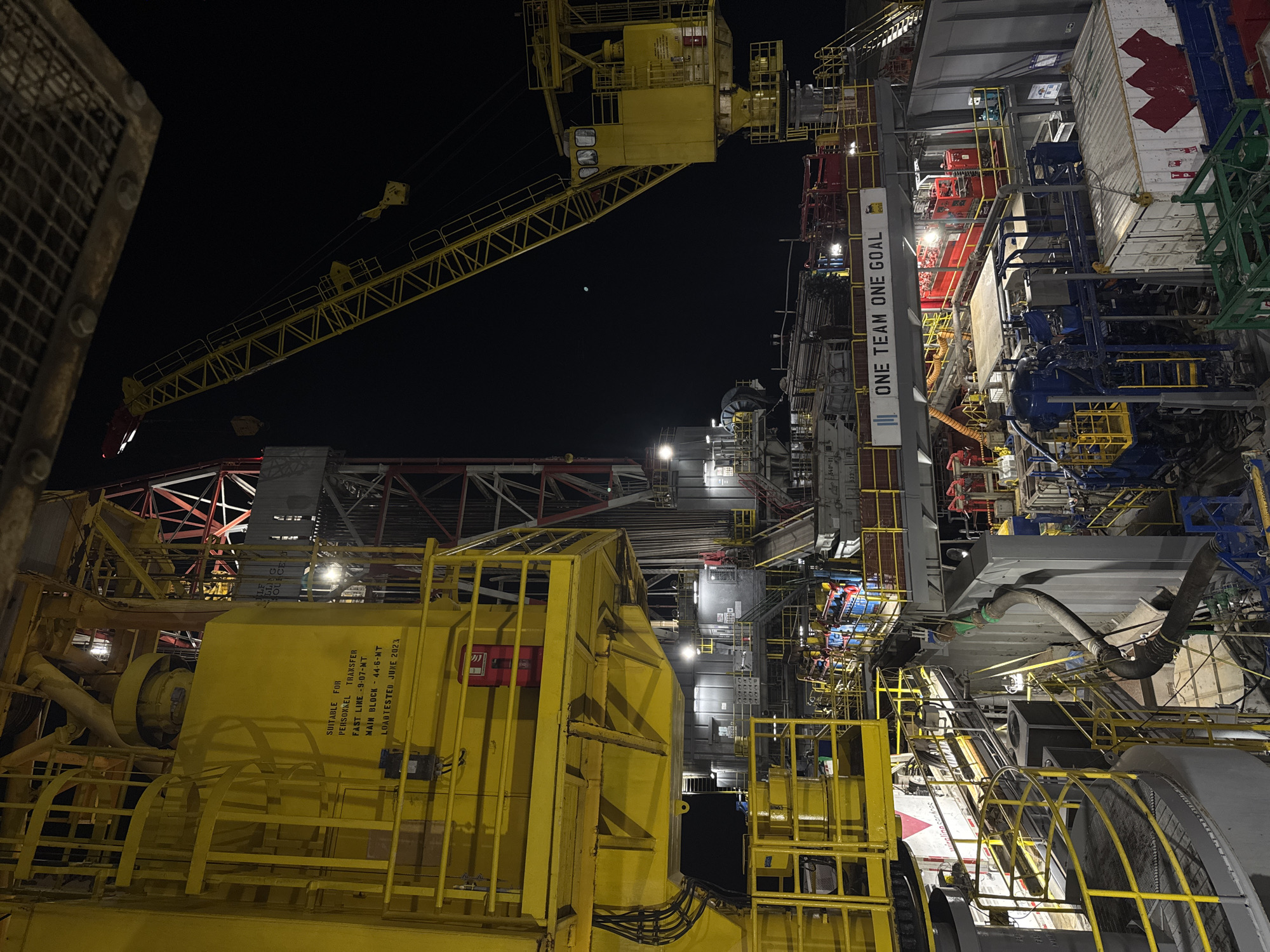 The image depicts an industrial offshore drilling rig at night. The scene is dominated by large yellow structures, intricate metal framework, and various pieces of heavy machinery. A large yellow crane extends from the right side of the frame, its boom silhouetted against the dark sky. Several levels of platforms and walkways are visible, some with yellow railings and stairs. Multiple lights illuminate different parts of the rig, creating strong contrasts and shadows. In the foreground, complex machinery with blue and gray components is present, along with large hoses and cables. A white shipping container with a red maple leaf symbol is visible on the lower right. A banner with the text "ONE TEAM ONE GOAL" is mounted on an elevated walkway. No people are clearly discernible in the image. The overall environment appears to be a dark, operational industrial site, likely at sea given the nature of the equipment. The location is specified as Unknown, Italy.
