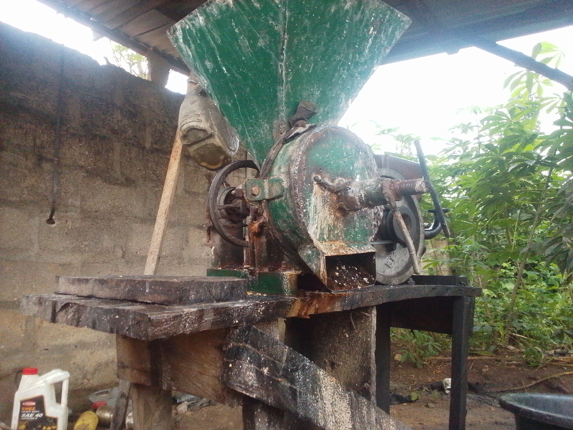 The image depicts a rustic, outdoor setup in Serti, Nigeria, likely a workshop or processing area. In the foreground, a weathered, dark wooden table supports a large, green, industrial grinding machine. The machine appears old and worn, with visible rust and residue. A funnel-like hopper at the top is filled with what seems to be processed material, perhaps grains or seeds, which are also seen spilling out. A large wheel, part of the machine's mechanism, is visible on the left.
The table itself is constructed from rough-hewn wooden planks, showing signs of age and use. Below the table, a support structure is made of similar weathered wood. To the left of the table, a white plastic bottle with a red cap, possibly containing oil, is placed on the ground. The background consists of a rough, concrete-block wall, partially covered by a wooden plank leaning against it. Beyond the wall, lush green vegetation thrives, suggesting a tropical or sub-tropical environment. The sky is bright and overcast, indicating daylight. The overall scene conveys a sense of manual labor and agricultural processing in a simple, unrefined setting.