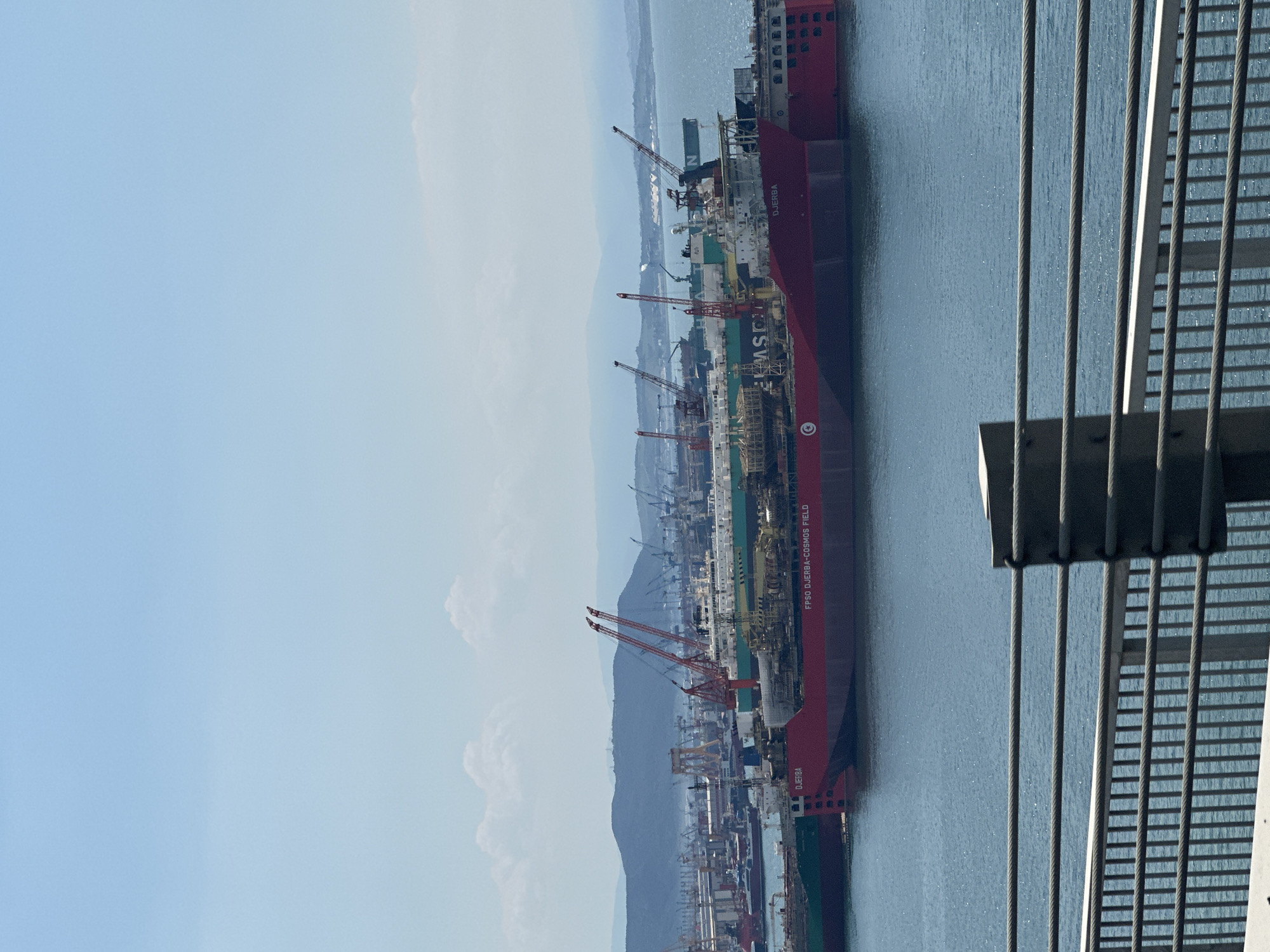 The image shows a large ship in a port. The ship has the name "DJERBA" and "FPSO DJERBA-COSMOS FIELD" on it. There are numerous cranes and other industrial equipment surrounding the ship, suggesting it's a shipyard or port facility. In the background, there are hills and a hazy sky. The photograph appears to have been taken from an elevated position, possibly from a bridge or building, looking down at the port. In the foreground, there are architectural details of a structure such as a bridge or an edge of a building with steel cables. Given the information about Altınova, Turkey, it is likely the ship is located at one of the shipyards in that area.