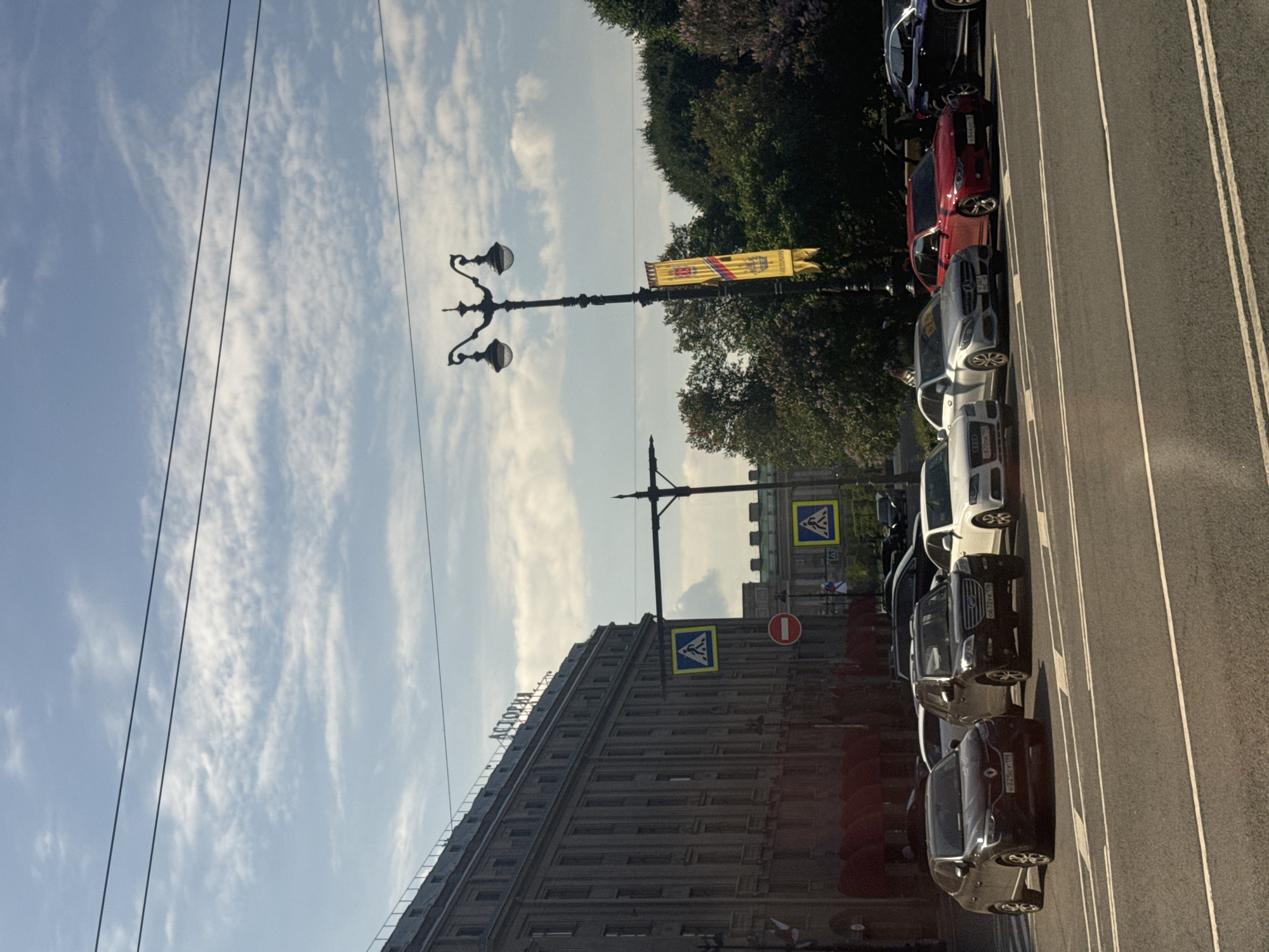 The image shows a street scene in Saint Petersburg, Russia, likely on Nevsky Prospekt, in front of the Astoria Hotel. The Astoria Hotel, a grand, multi-story building with a classical facade and many windows, dominates the left side of the frame. The hotel sign, "АСТОРИЯ" (Astoria), is visible near the roofline. A pedestrian crossing sign with a blue square and a white pictogram of two figures walking is positioned on the sidewalk in front of the hotel. Red awnings are visible at the base of the building.
The street itself is paved and has white lane markings. Several cars are parked or driving on the street. These include a dark grey Renault sedan in the foreground, a dark SUV, a white Audi sedan, a silver Mercedes-Benz sedan, a red BMW sedan, and a dark blue SUV. A streetlamp with two ornate globes stands on the right side of the street, with a yellow banner displaying some sort of insignia hanging from it. In the background, lush green trees and some flowering bushes are visible, along with other buildings and a clear blue sky with scattered clouds. Another pedestrian crossing sign is visible further down the street, as well as a red circular sign with a white horizontal bar, indicating a prohibition. The overall scene suggests a sunny day in a bustling urban environment.