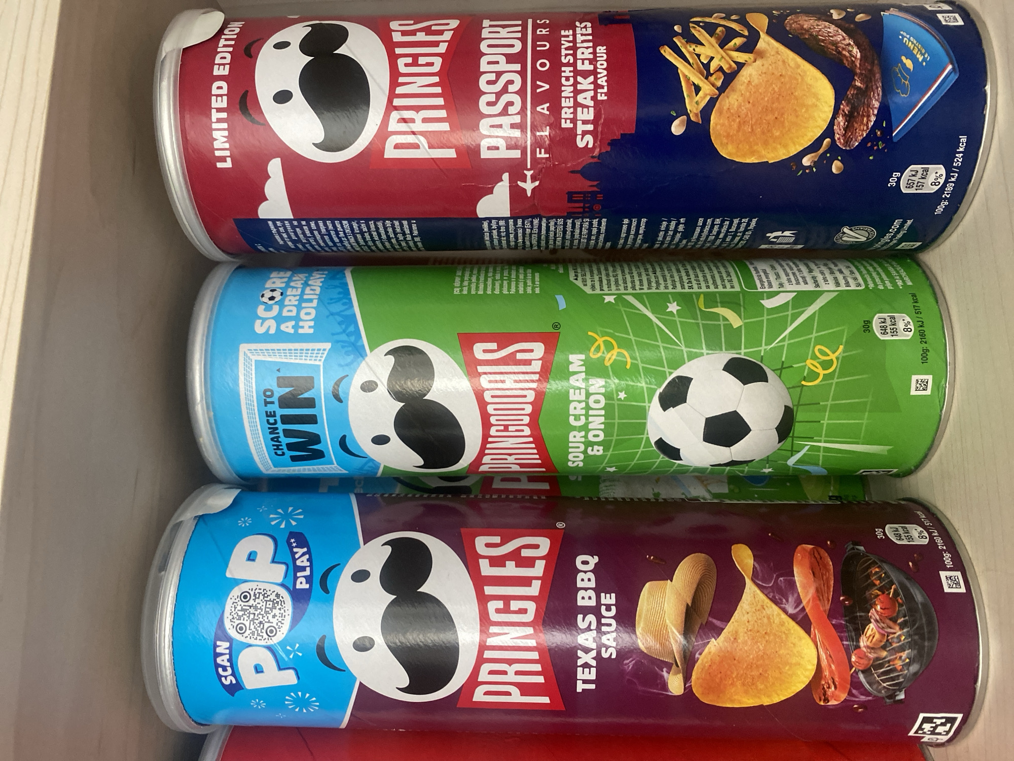 The image shows three Pringles cans on a shelf. The Pringles cans are purple, green, and red. The purple can has "Pringles Texas BBQ Sauce" written on it. The green can has "Pringles Sour Cream & Onion" written on it and features a soccer ball. The red can has "Pringles Passport Flavours French Style Steak Frites" written on it and shows a cityscape. The image is taken from a medium shot, focusing on the cans. The background is blurred, but appears to be a store shelf. The lighting is bright and even. There are no people or animals visible in the image. The location is not specified in the image, and therefore cannot be determined to be Chișinău, Moldova.