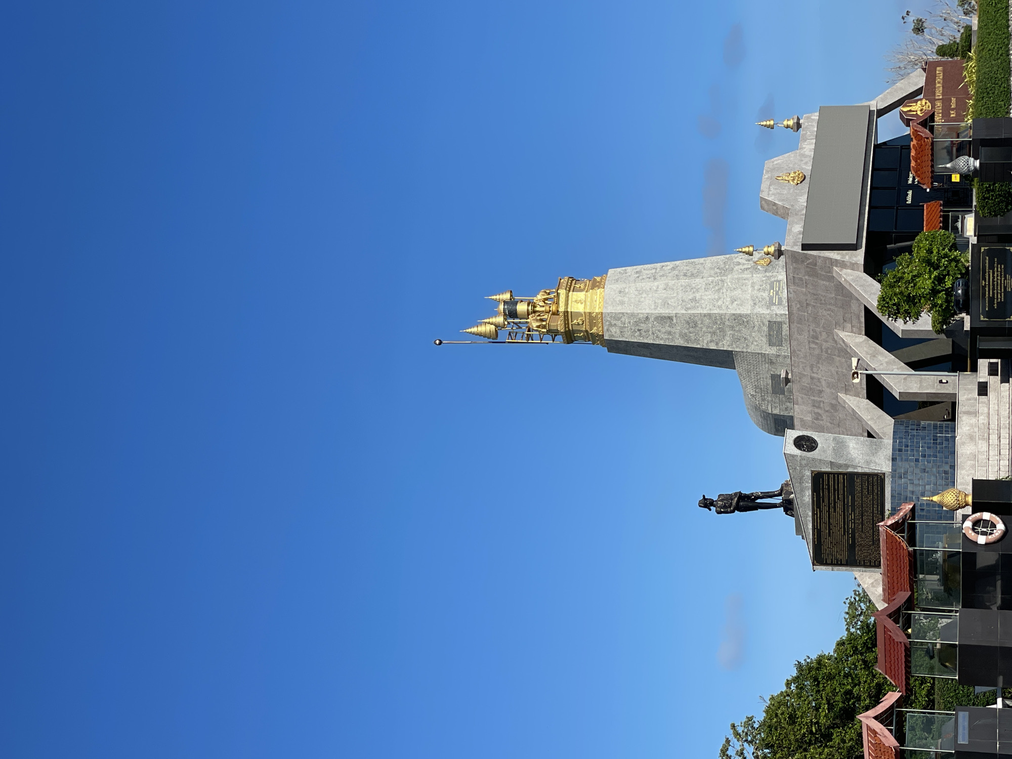 The image shows the Promthep Cape monument in Rawai, Thailand, under a clear blue sky. The monument is a large, multi-tiered structure made of grey stone, topped with a golden stupa-like spire adorned with intricate carvings and statues. A dark bronze statue of a man in a military uniform stands on a pedestal to the left of the main structure. In front of the monument, there are several covered display areas with red roofs and glass enclosures, showcasing what appear to be artifacts or models. A life preserver ring is visible near the base of the monument. Lush green trees and vegetation are present on the left and right sides of the scene. The overall impression is that of a historical or cultural landmark, likely a tourist attraction.