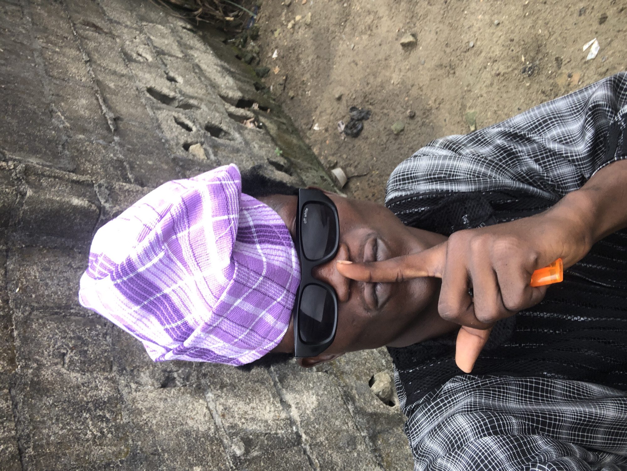 A young Black man, wearing a purple plaid cap, black sunglasses, and a black and white checkered shirt over a black mesh undergarment, stands in front of a rough, grey concrete wall. He is holding an orange lighter and has his index finger to his lips in a shushing gesture. The ground is sandy and uneven, with scattered debris. The scene appears to be outdoors, possibly in Ado, Nigeria, given the architecture and environment.