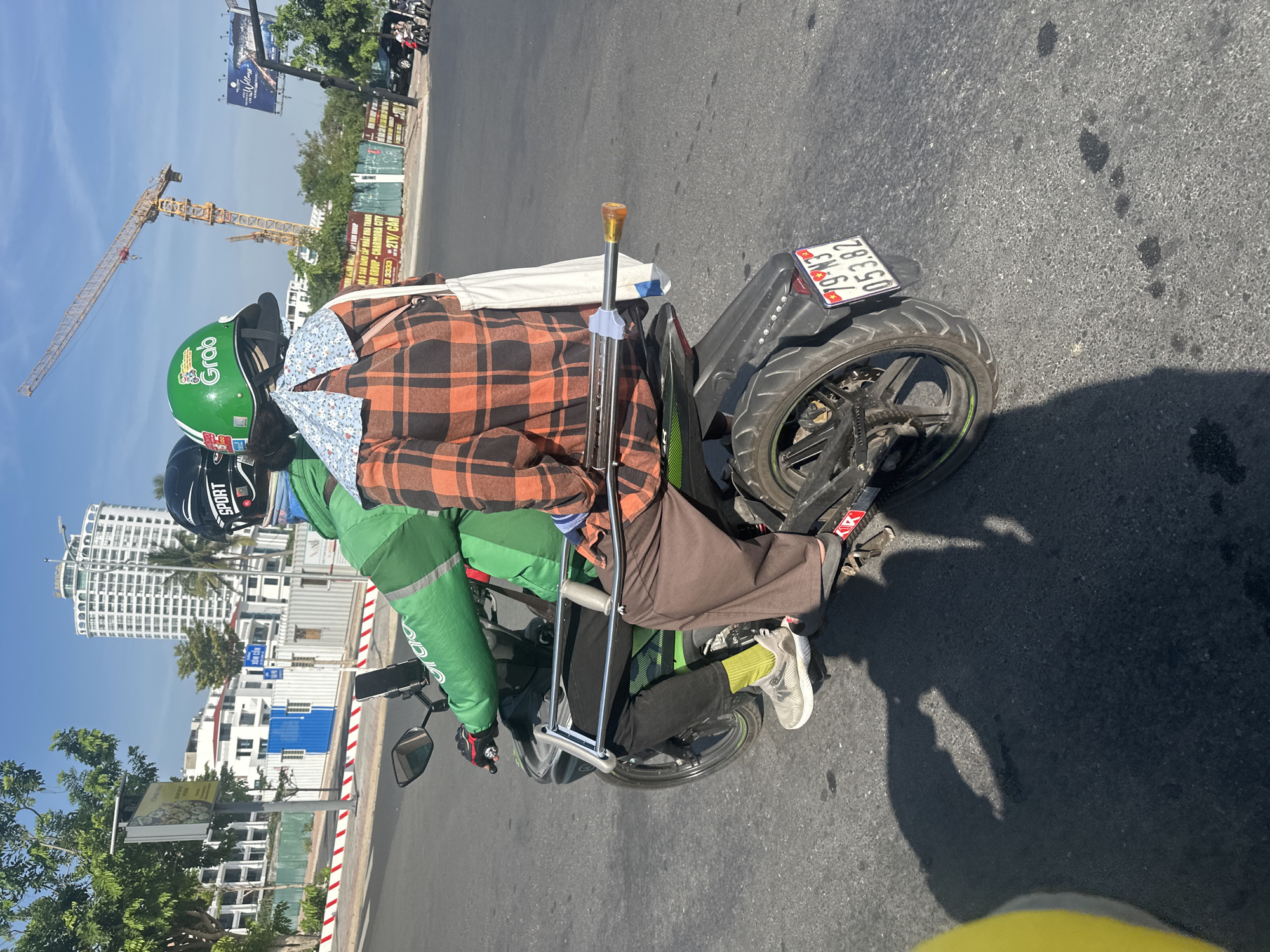 In Nha Trang Ward, Vietnam, two individuals are riding on a motorcycle. The person in the front, wearing a green jacket and a helmet with "SPORT" visible, is operating the vehicle. They have a phone mounted on the handlebars. The passenger in the back is wearing an orange and black plaid shirt and a blue and white floral neck scarf. They are holding a pair of crutches, which are positioned between them and the driver, extending from their lap to behind the motorcycle seat. The crutches have metal shafts and a light-colored handle. A white tote bag is slung over the passenger's shoulder. The motorcycle has a license plate with the numbers "79-N3 053.82". In the background, there is a construction crane, several multi-story buildings, and trees. The scene is illuminated by sunlight, casting shadows on the asphalt road.
