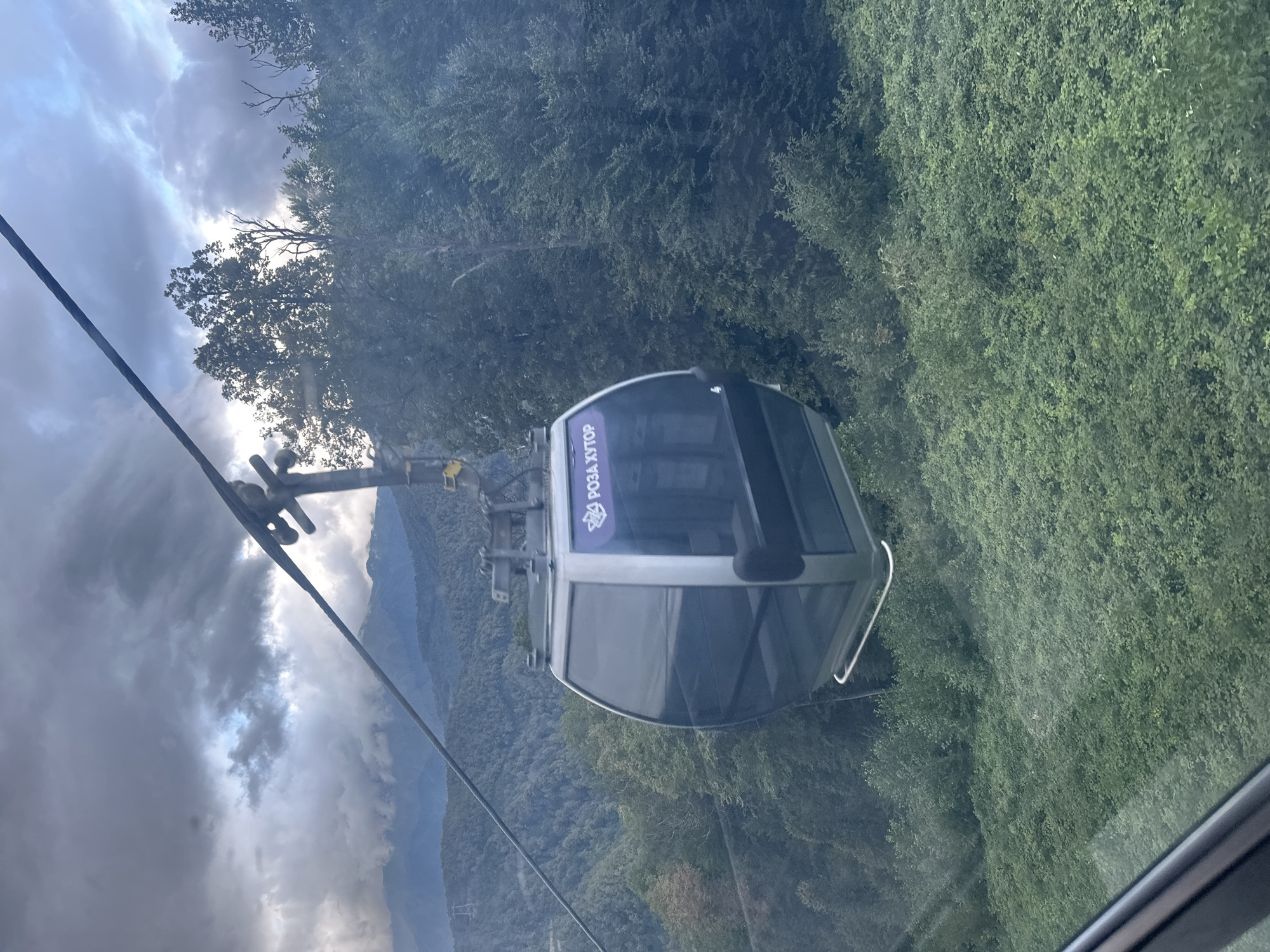 This image captures a cable car cabin ascending a mountainous incline in Estosadok, Russia. The cabin, primarily silver and black with dark tinted windows, features a purple logo with text that appears to be in Cyrillic. The cable car is suspended from a thick cable that stretches diagonally across the frame against a backdrop of dramatic, cloudy skies and distant, mist-covered mountains. The immediate surroundings are densely covered with lush green trees and foliage, indicating a natural, possibly forested environment. The perspective suggests the photograph was taken from another moving cable car or a similar vantage point, looking slightly upwards and towards the right. The lighting is somewhat dim, suggesting either an overcast day or early morning/late evening.