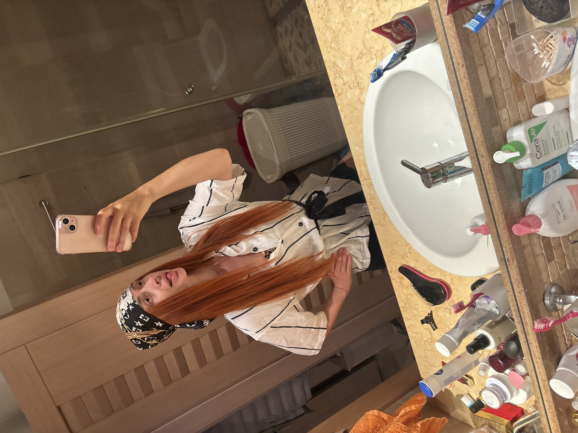 The media file shows a young woman with long, bright red hair taking a mirror selfie in a bathroom. She is wearing a white shirt with black pinstripes and a black and white bandana. The bathroom appears to be well-lit, and the time of day is not discernible. The setting includes a sink with a chrome faucet, a countertop with various toiletries including toothbrushes, lotions, and other personal care items. A laundry basket is visible in the background. The woman is sticking her tongue out, suggesting a playful or lighthearted mood. The phone she is holding is a light pink iPhone. The image is taken from a slightly high angle, looking down on the woman. The background is slightly blurred, focusing attention on the woman and the immediate area around the sink. The overall impression is a casual snapshot taken in a domestic setting. The location context of Budva, Montenegro is not visually evident in the image.