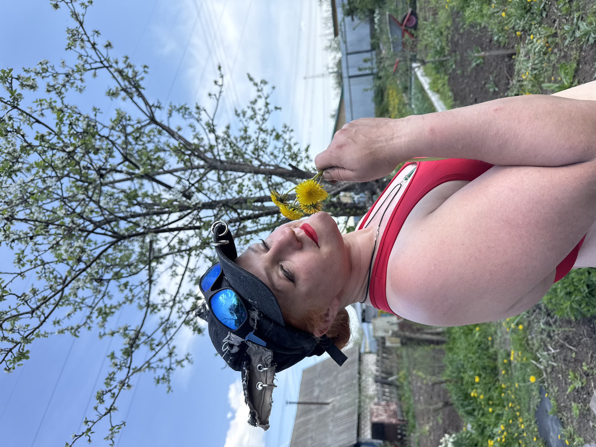 A woman is holding a bunch of yellow dandelions in an outdoor scene in Chaikovsky, Russia. The woman is wearing a red top, a black hat adorned with goggles, and red lipstick. A blooming tree is on the left side of the frame against a blue sky with power lines visible in the background. In the background are parts of buildings and a garden area with greenery. The image is oriented sideways.