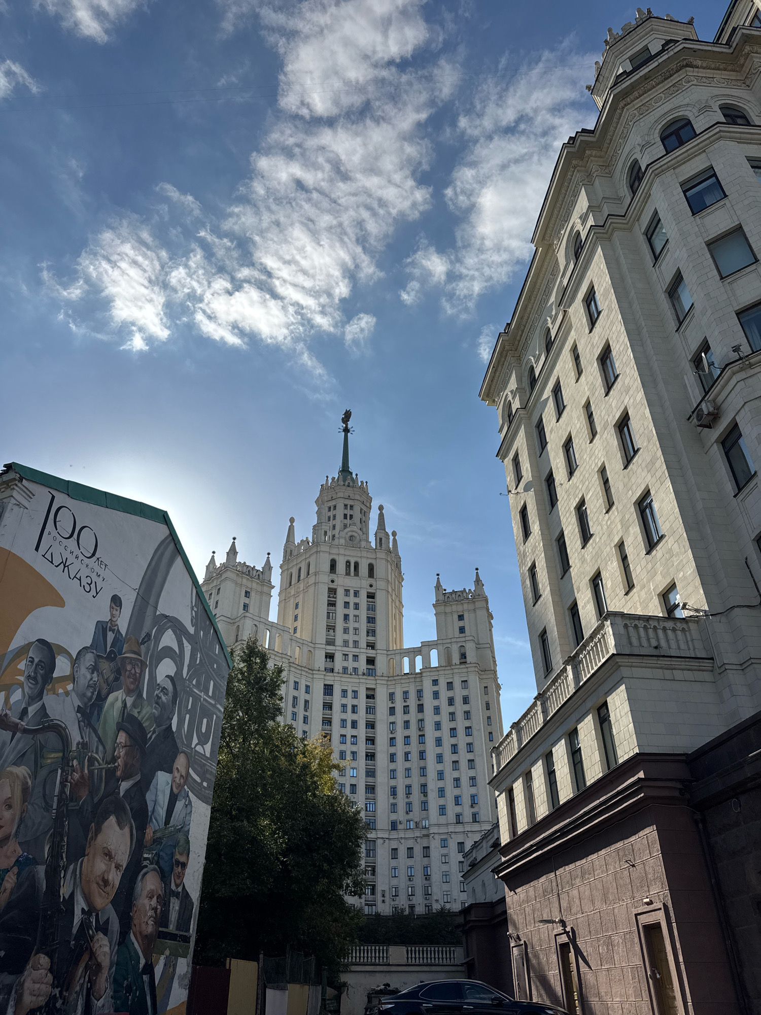 The image depicts a street scene in Moscow, Russia, featuring distinct architectural styles and a mural. In the foreground, a mural is visible on the side of a building, depicting portraits of several men and the text "100 лет Российскому Джазу" (100 years of Russian Jazz). The mural is vibrant and detailed, showcasing individuals in formal attire, some holding musical instruments.
Behind the mural, two prominent Stalinist skyscrapers dominate the skyline. The skyscraper on the right is a large, cream-colored building with numerous windows arranged in a grid pattern, and ornate architectural detailing at its corners and roofline. It has several projecting sections and balconies. To the left of this, a slightly smaller but equally grand Stalinist skyscraper is visible. This building also features a central spire topped with a sculpture and decorative elements, with multiple levels and a symmetrical design.
The sky above is a bright blue with scattered white clouds, indicating daytime. A faint sun is visible behind the clouds, casting some glare. The overall environment suggests an urban setting with significant historical architecture. A dark-colored car is parked on the street in the lower right portion of the image. Trees are present between the buildings, contributing to the urban greenery. The perspective of the shot is from a low angle, looking upwards towards the imposing structures.