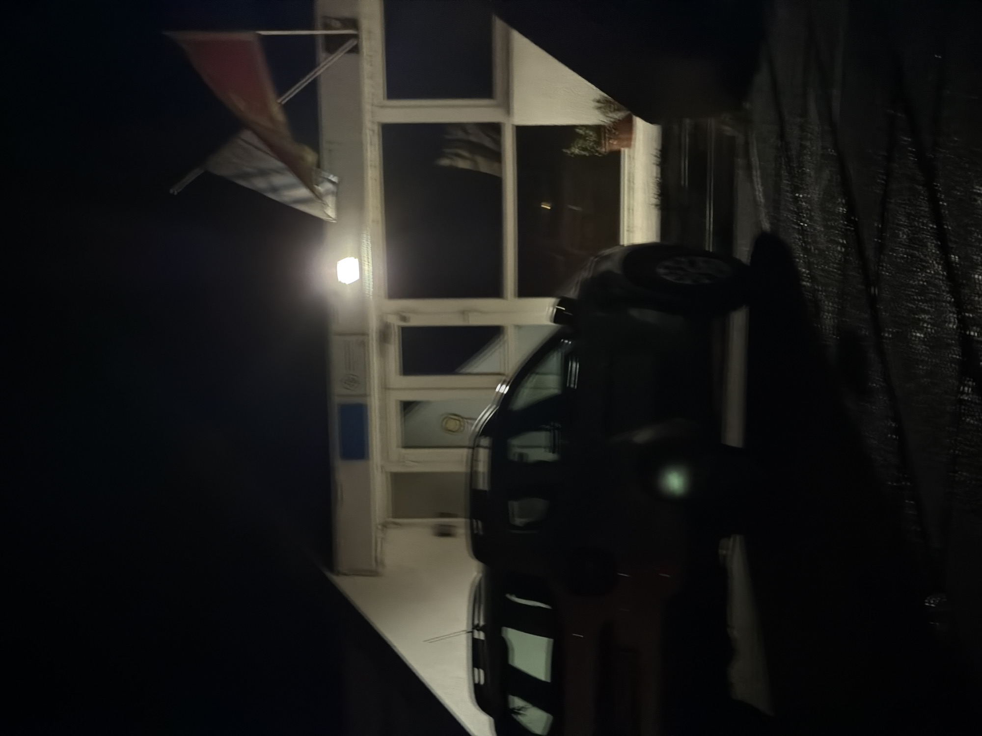 The image depicts a nighttime scene at an exterior location in Podgorica, Montenegro. A vehicle is partially visible on the right side of the frame. In the background, a building is present with windows and a flag mounted on its exterior. A light fixture illuminates the building. The environment is dark, with limited ambient lighting.