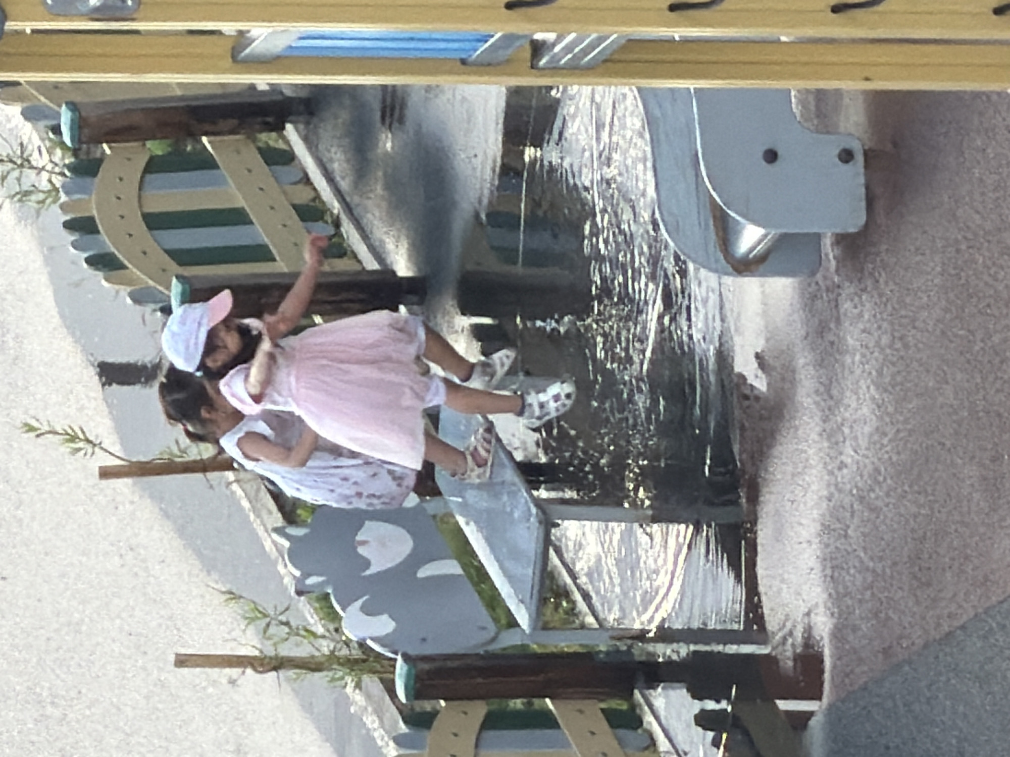 The image shows two young girls on a wet playground. One girl, wearing a white dress, stands on a metal bench. The other girl, in a pink dress and a white and pink cap, is stepping off the bench. The playground appears to be wet, with puddles of water on the ground, suggesting it may have rained recently. The background shows a fence with colored wooden slats and some green foliage. The image was taken from an elevated angle. The style of the playground equipment and the presence of vegetation could be consistent with a park or recreational area in Astana, Kazakhstan, known for its modern infrastructure and public spaces.