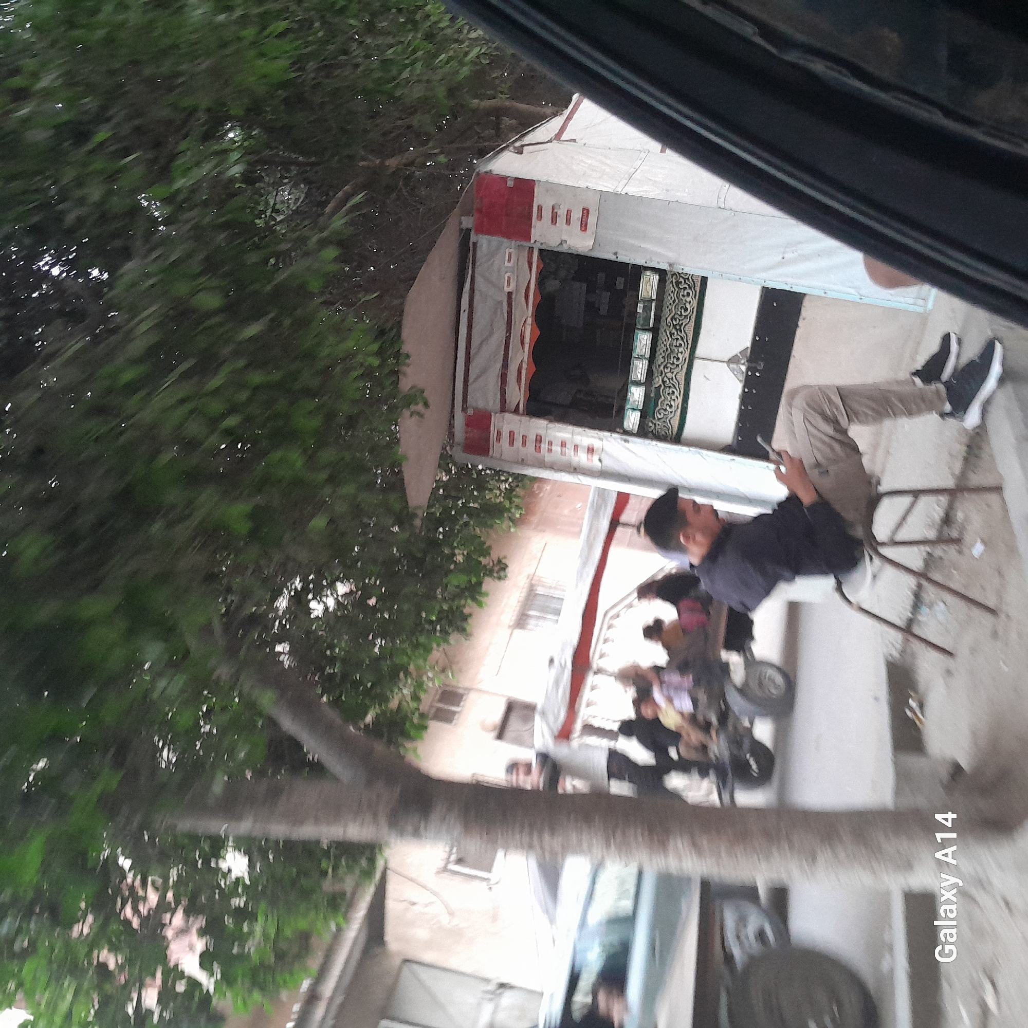 In Khan Yunis, Palestinian Territories, a man sits on a chair in front of a roadside stall, engrossed in his phone. The stall, draped in white and adorned with red accents and patterned fabric, appears to be selling goods. Behind the stall, a cart with two wheels is visible, carrying people. A car is parked to the left of the scene, and a large tree with lush green foliage provides shade. The environment suggests a typical street in a populated area, with buildings visible in the background. The overall impression is one of everyday life unfolding in a commercial district.