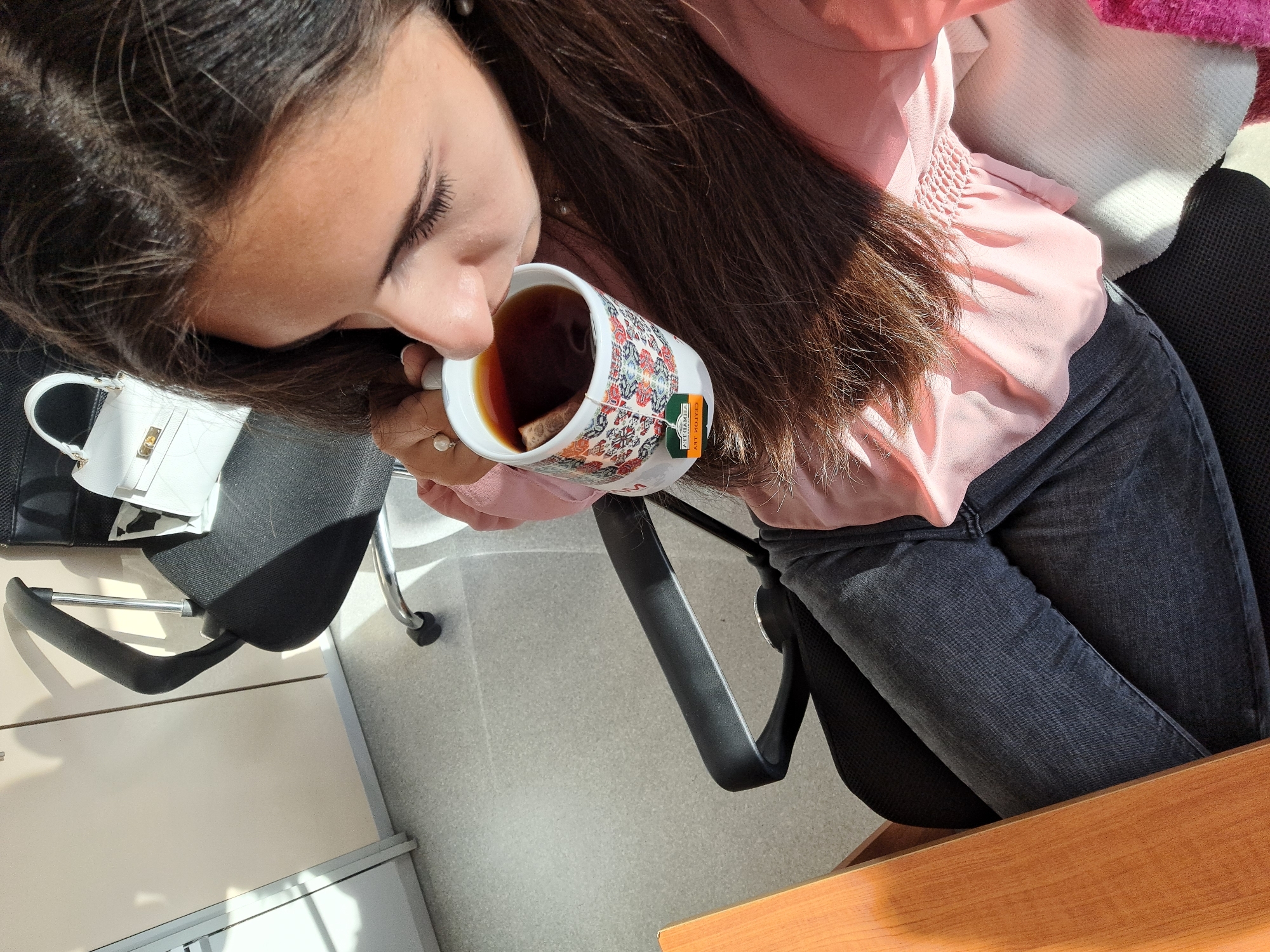 This high-angle image captures a person enjoying a cup of tea in an indoor setting, likely an office or study. The person, with long, dark brown hair, is positioned diagonally from the upper left to the lower right. They are wearing a light pink, possibly smocked, blouse and dark grey or black jeans.
Their head is tilted, bringing their nose close to a white ceramic mug, which they hold with their right hand. The mug features intricate, colorful patterns, resembling a folk or ethnic design. Inside, a dark brown liquid, presumably tea, contains a tea bag with a prominent green and orange tag. Visible text on the tag reads "CEYLON TEA" and "PURE CEYLON." The person's eyes are closed or cast downwards, suggesting a moment of relaxation, contemplation, or savoring the aroma and taste of the tea. A ring with a pearl or light-colored stone is visible on their right ring finger.
To the upper left, a small, white, quilted-pattern handbag with a gold clasp rests on a black office chair. Portions of other black office chairs are also visible. Strong, natural sunlight streams into the scene, casting distinct shadows on the light grey floor and a light wooden desk or table surface in the lower right foreground. The bright lighting indicates it is daytime. The overall scene depicts a peaceful and introspective moment, possibly a tea break.