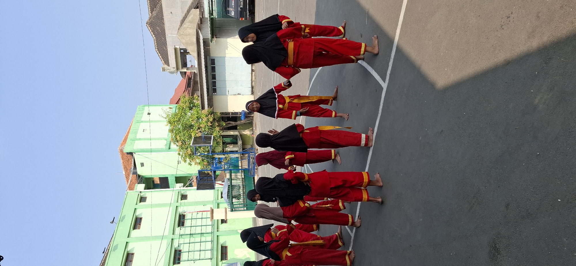 In the city of Wonorejo, Indonesia, a group of children, likely female students, are gathered in an outdoor paved area, possibly a schoolyard or sports court. They are dressed in traditional red martial arts uniforms with yellow accents, and most are wearing black or colored hijabs. The children appear to be in the middle of a training session or a break, with some standing and others interacting. The environment includes a green building in the background, a basketball hoop, and trees. The sky is clear and blue, indicating a sunny day. The scene is captured from a slightly elevated perspective, looking down at the group.