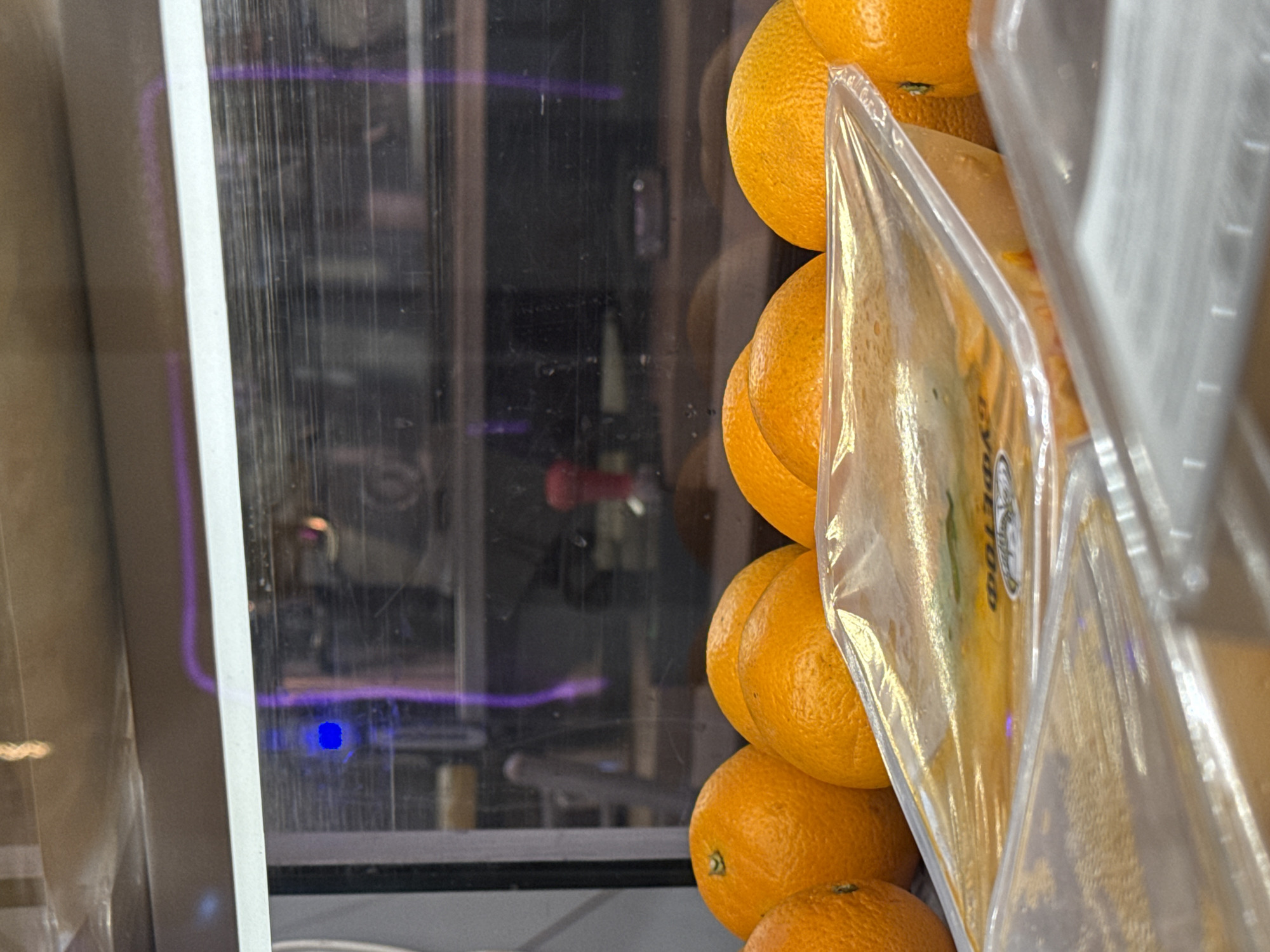 The image depicts a close-up of stacked oranges in a refrigerated display case. The oranges are bright orange with a textured peel and are packaged in clear plastic wrap. The display case is made of metal and glass with visible streaks, fingerprints, and condensation. A faint blue light is visible on the inside of the display. The specific location in Moscow, Russia, is not discernible from the image. No people are visible in the scene.