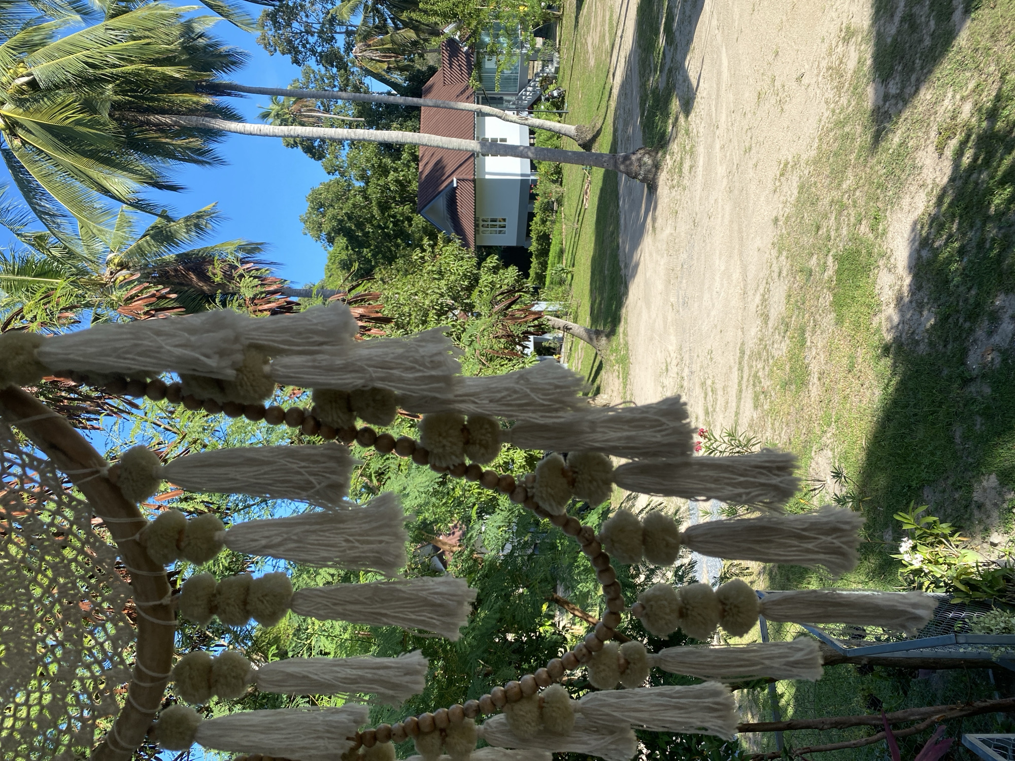 The image depicts a scene in Ko Pha Ngan, Thailand. In the foreground, a handmade macrame decoration with white tassels and wooden beads hangs from a branch, obscuring part of the view. Beyond the decoration, a white house with a reddish-brown roof is visible, set amidst lush green trees and palm trees. The ground in front of the house is a mix of sand and grass. A clear blue sky is visible above the trees. The overall impression is one of a tropical, serene environment, possibly a resort or a private residence in a tropical location.