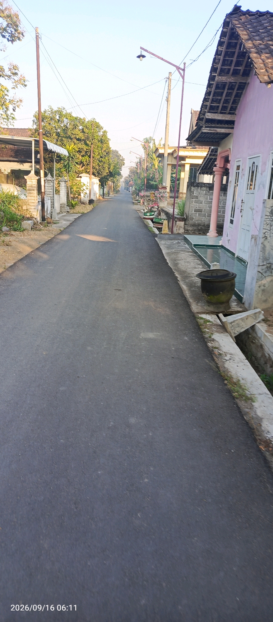 The image shows a straight, paved road in Karangsono, Indonesia. The road is lined with houses on both sides. The house on the right is painted pink and has a tiled roof. There are decorative pillars and fencing in front of the houses. A large clay pot is visible on the right side of the road, near the house. The road appears to be in a residential area, with trees and vegetation also present. The sky is clear and a street light is visible in the distance. The timestamp "2026/09/16 06:11" is present in the bottom left corner of the image.
