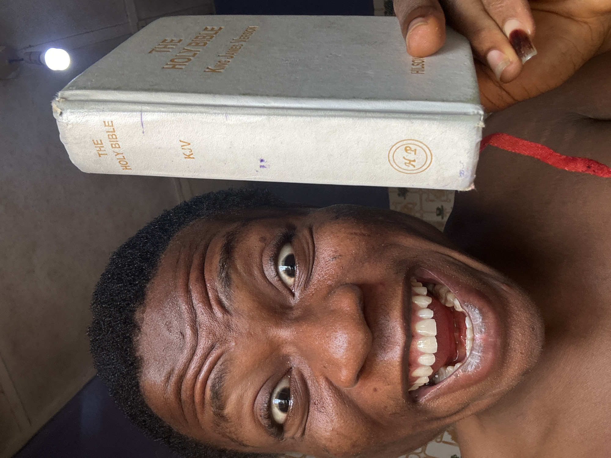 A close-up, upside-down, eye-level shot features a person with brown skin holding a white hardcover King James Version of the Holy Bible on their head. The person is smiling widely, exposing upper and lower teeth. Wrinkles are visible on their forehead. The location is Unknown, Unknown.