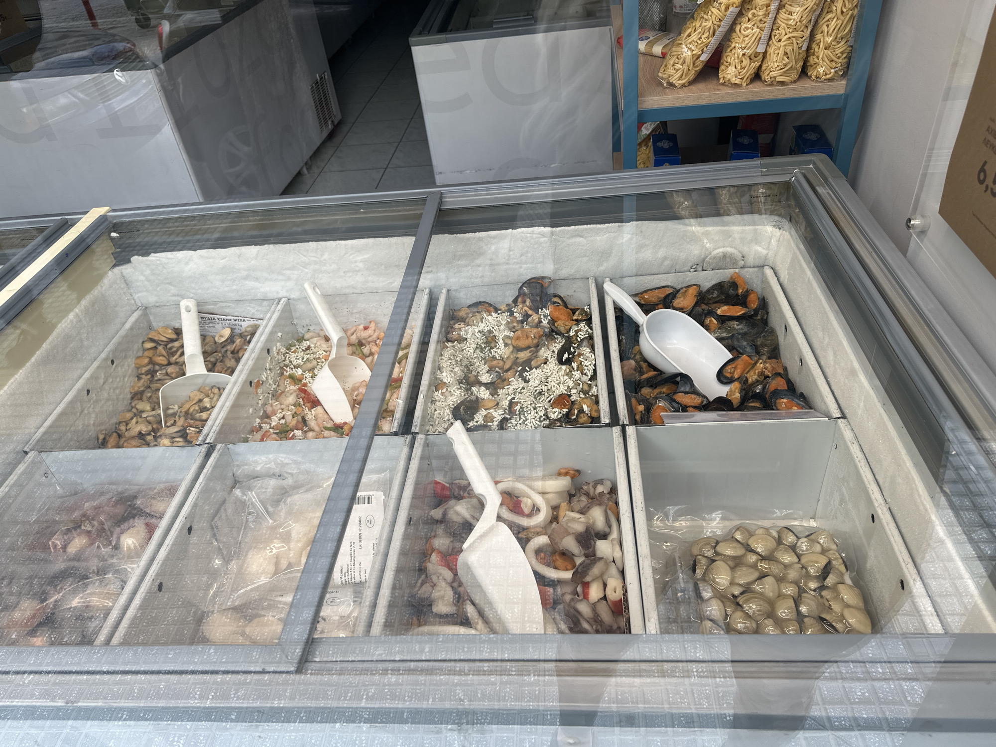 The image shows a freezer display case in a store, likely located in Serres, Greece. The display case contains various types of frozen seafood, including mussels, clams, shrimp, and squid. Each type of seafood is in its own compartment and most have a plastic scoop in them. The freezer case is clean and well-lit. A shelf of pasta is visible behind the freezer.