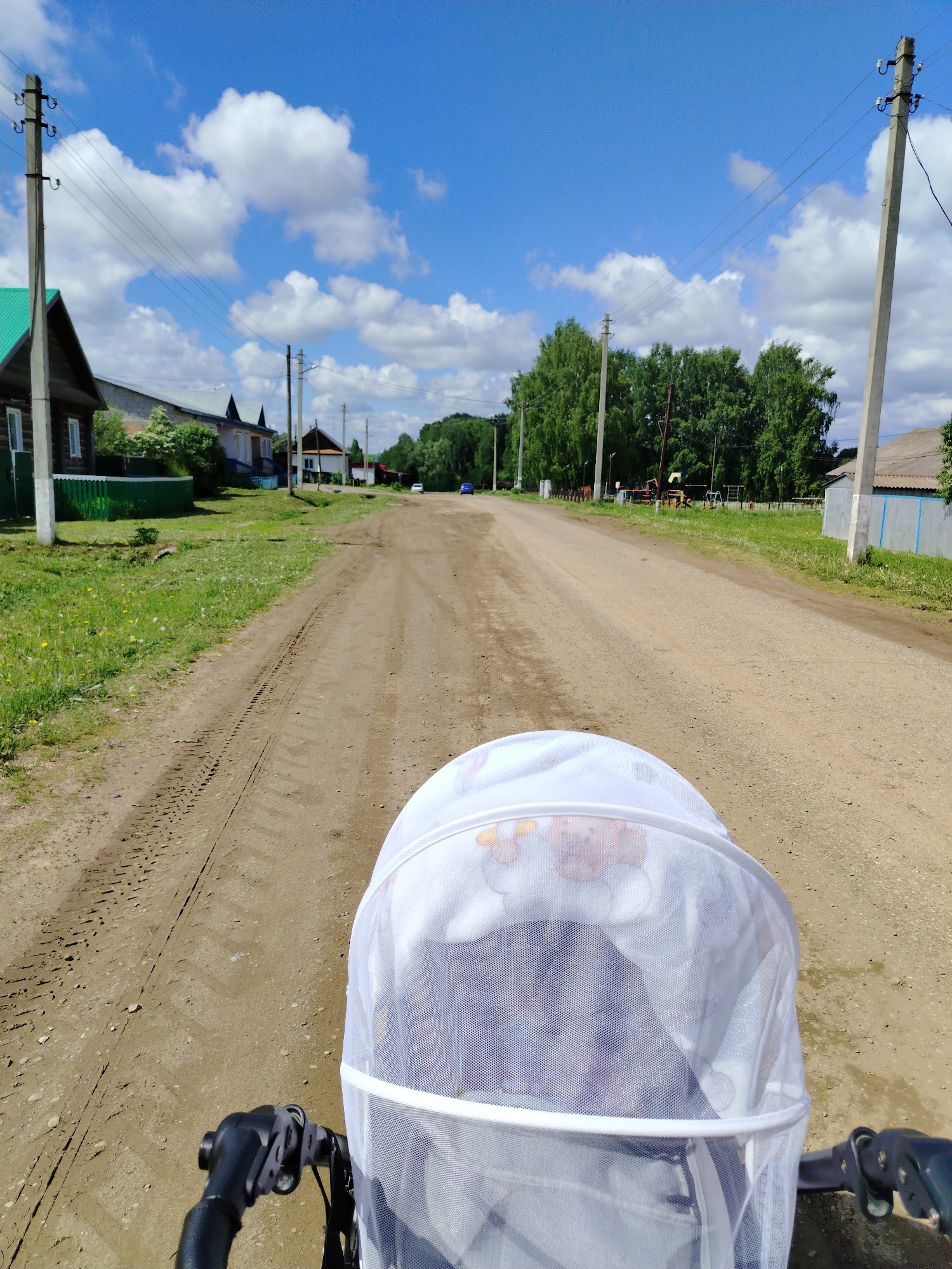 A ground-level shot shows a dirt road stretching through what appears to be a residential area, likely within the city of Гожан, Russia, given the query context. The foreground is dominated by a baby stroller's hood, its white mesh cover suggesting it's in use. The stroller's handlebars are visible at the lower edges of the frame. The road itself bears tire tracks, indicating vehicle traffic. On either side of the road are houses, some with green roofs, interspersed with trees and power lines. The sky is bright blue, dotted with cumulus clouds, suggesting a sunny day.