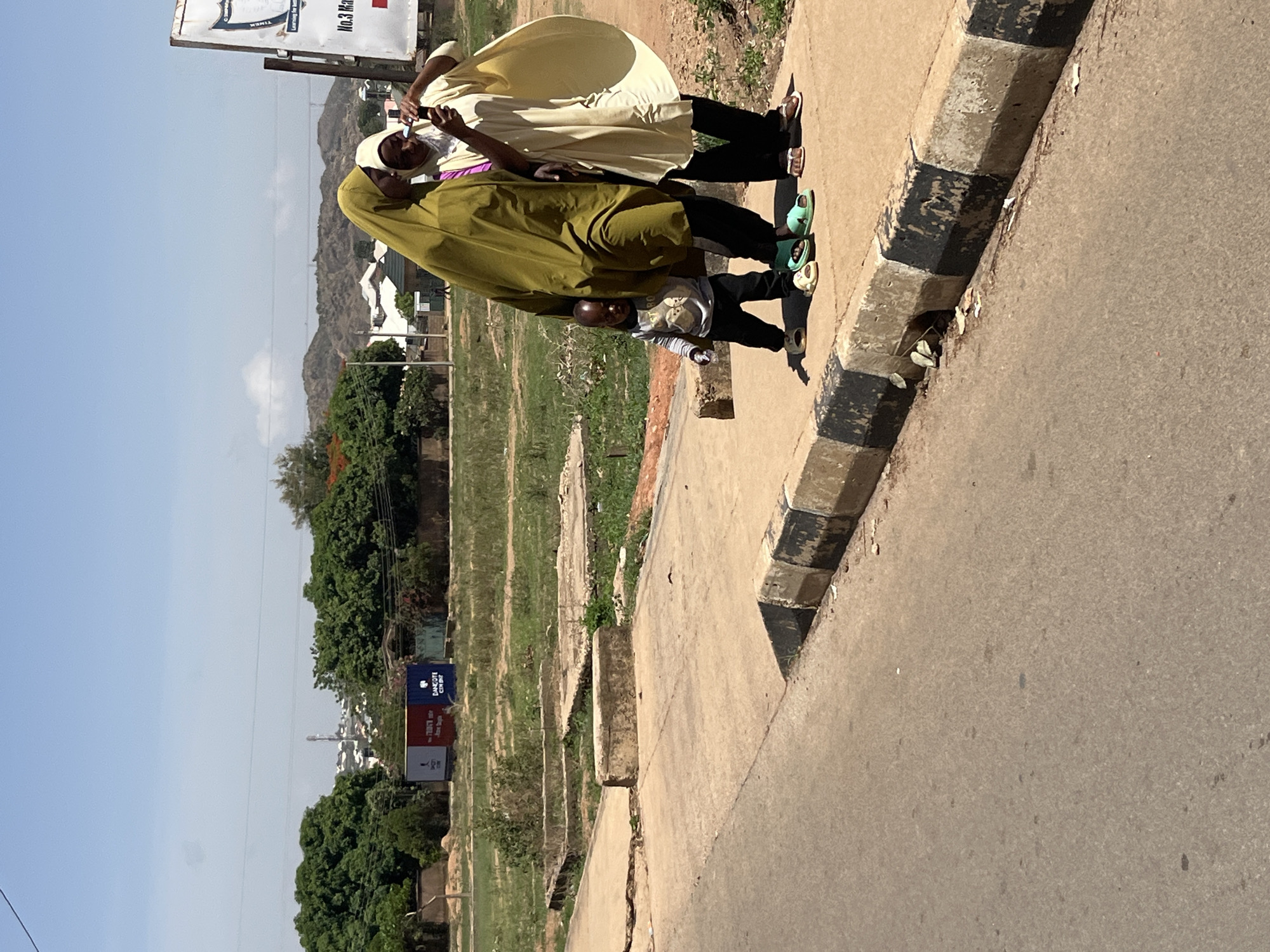 Three people are walking on a sidewalk next to a road. One person is wearing a light yellow robe, another is wearing a green robe and the third is a child. The first two people are wearing white head coverings. The road has a black and white curb. In the background is a hillside and some storage containers. The image was taken in Gura Top, Nigeria.