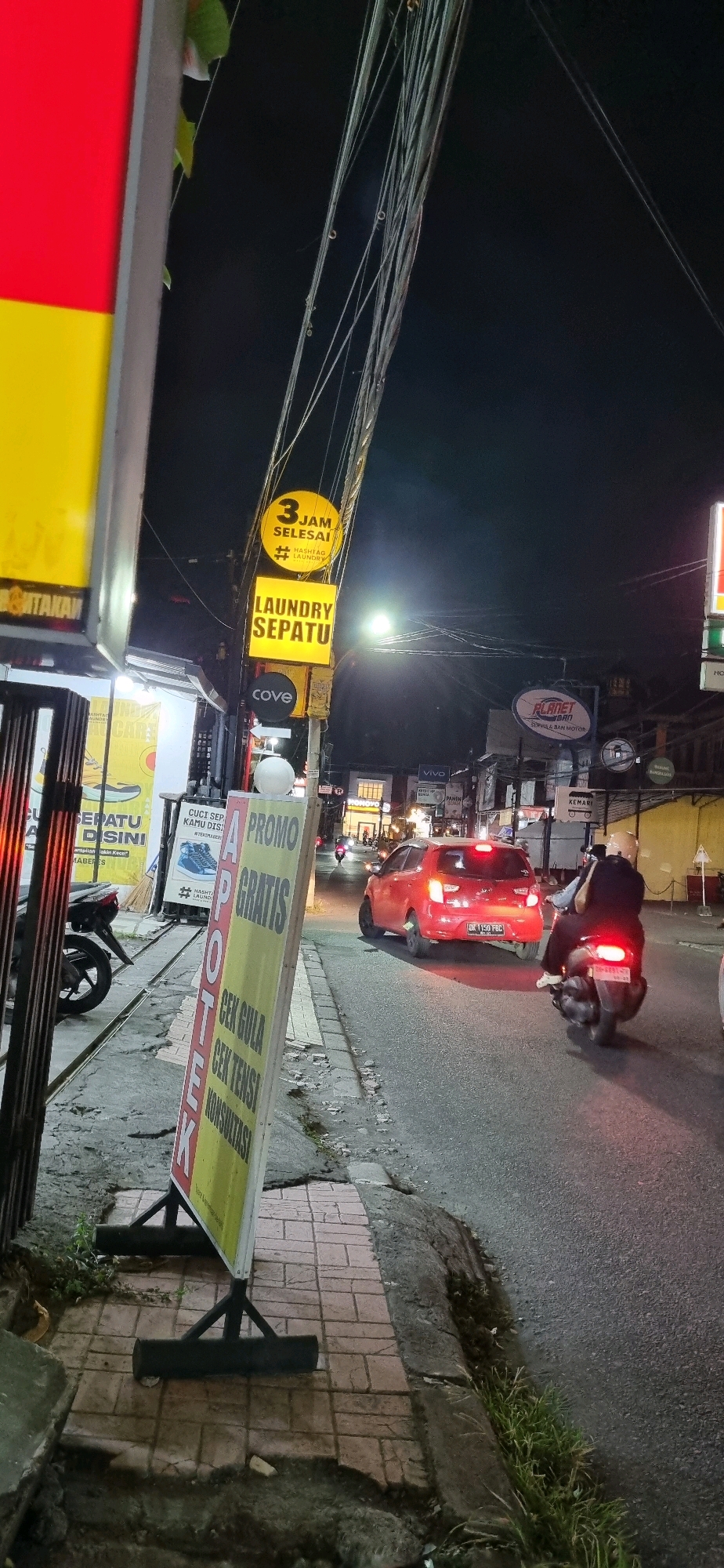 The image depicts a street scene at night in Denpasar, Indonesia. A red car is visible driving on the right side of the road, with its brake lights illuminated. A scooter with two occupants is also on the road, following the car. The street is lined with various businesses and signage.
To the left, there is a tall, narrow sign with red and yellow panels. Further down the sidewalk, an A-frame sign for "APOTEK" (pharmacy) is displayed, with text indicating "PROMO GRATIS" (free promo), "CEK GULA" (check sugar), "CEK TENSI" (check blood pressure), and "KONSULTASI" (consultation). Adjacent to this sign is another sign for "LAUNDRY SEPATU" (shoe laundry) with a "3 JAM SELESAI" (3 hours done) emblem.
Overhead, a complex network of electrical wires is visible against the dark night sky. In the background, more illuminated signs for businesses are present, including one that reads "VIVO". The ground is paved with bricks on the sidewalk and asphalt on the road, with some areas showing signs of wear and tear. Small patches of grass are growing along the edge of the road.