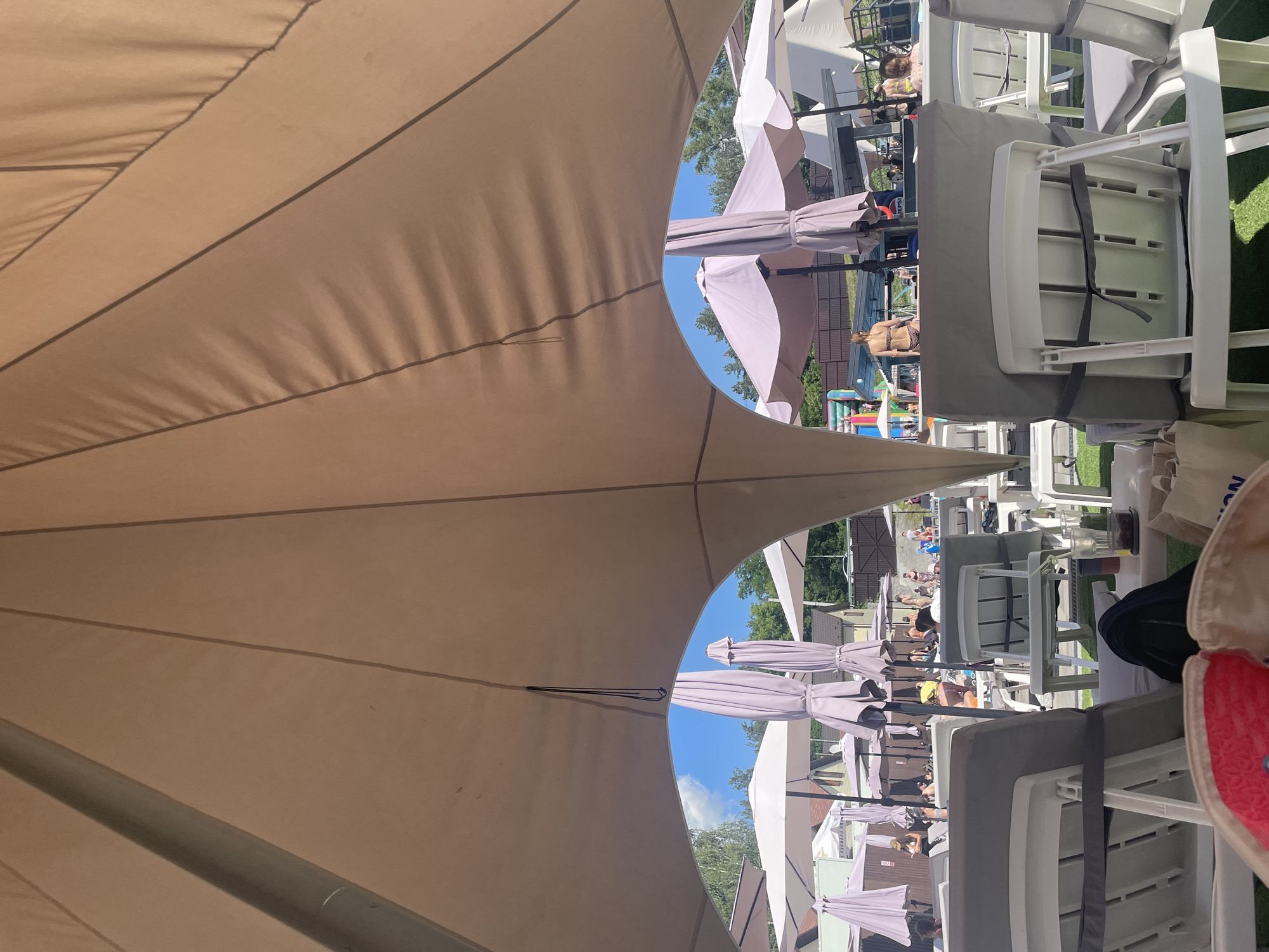 The image depicts an outdoor area with lounge chairs and umbrellas, suggesting a resort or recreational facility. The foreground features several white and grey lounge chairs covered by a large, beige, tent-like canopy, providing shade. In the background, under a clear blue sky with scattered clouds, more umbrellas and lounge chairs are visible. People are scattered throughout the area, some seated on chairs and others standing. Some individuals are dressed in swimwear, indicating a poolside or beach environment. Buildings, trees, and a playground structure are also present in the distance. The scene appears to be a sunny day, with artificial light sources not clearly visible. The location is identified as Zazymia, Ukraine, suggesting a summer scene at a place like the "Zazymia Resort and Spa".
