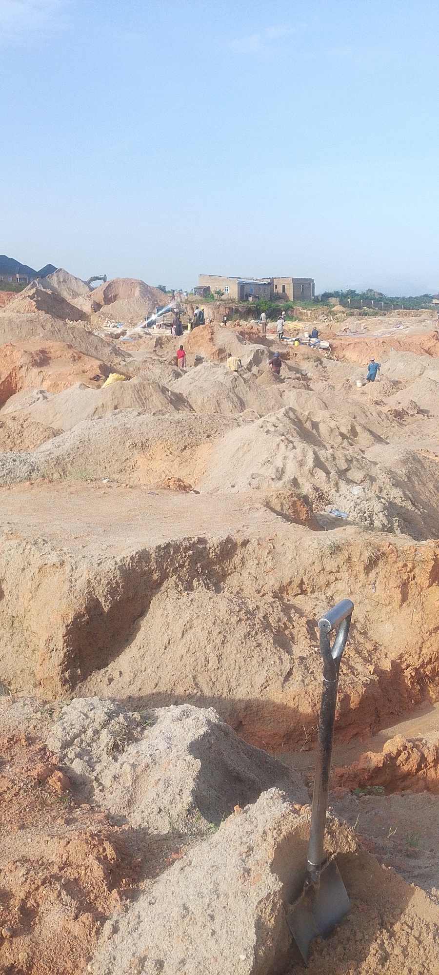 The image depicts an active construction or excavation site in Rayfield, Nigeria, under a clear, pale blue sky. The landscape is dominated by large mounds of sandy earth, varying in color from light tan to reddish-brown, indicating disturbed soil. In the foreground, a shovel with a dark handle is partially embedded in a pile of lighter sand.
Scattered throughout the site are numerous individuals, likely laborers, engaged in various activities. Some appear to be digging or shoveling, while others are standing or walking among the mounds. A few individuals are seen carrying or moving materials. One person in the mid-ground is wearing a red garment. In the background, several buildings are visible, suggesting a developing area. One larger, light-colored building with visible windows and doors appears to be under construction or recently completed. The overall environment is dry and dusty, consistent with a sand quarry or an area undergoing extensive earthmoving. There are no obvious signs of heavy machinery other than what might be a distant excavator partially obscured by a sand dune.