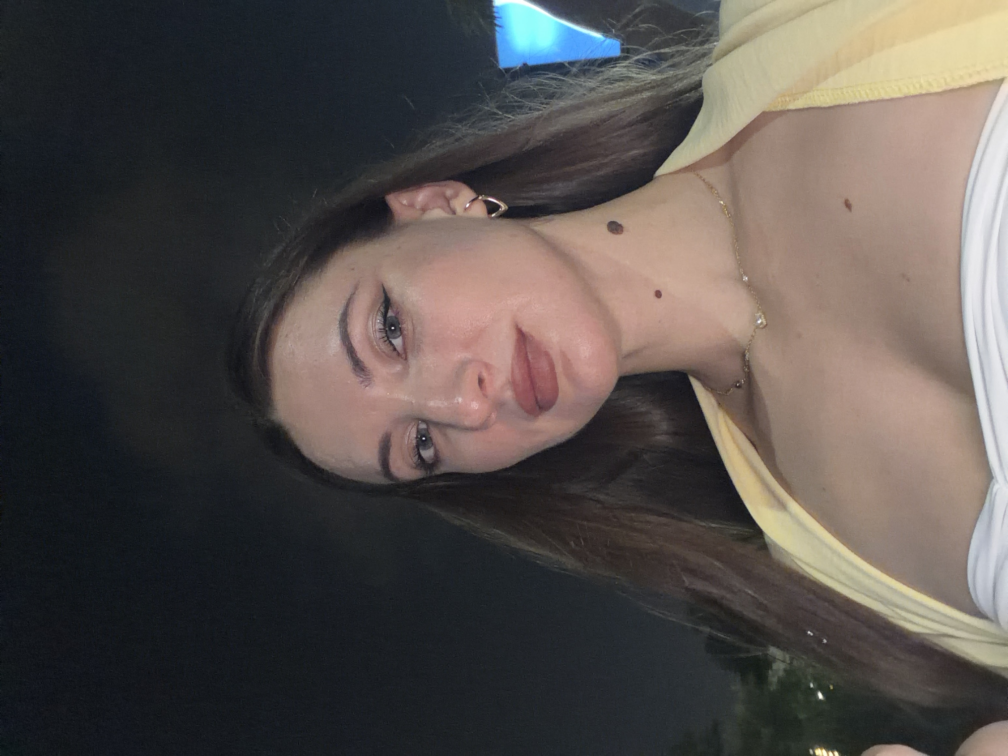 The image shows a close-up, selfie-style shot of a woman's face and upper torso. She has long, dark brown hair, fair skin, and is wearing makeup, including winged eyeliner and lipstick. She has a gold hoop earring in her left ear and a delicate gold necklace with small pendants. There are two moles visible on her neck, and one on her chest. She is wearing a white strapless top and a light yellow garment draped over her shoulders. The background is dark, suggesting it is nighttime, with some faint lights and shapes visible, including what appears to be a bright blue light on the right side. There is no specific indication of the location being the city of 6th of October, Egypt.