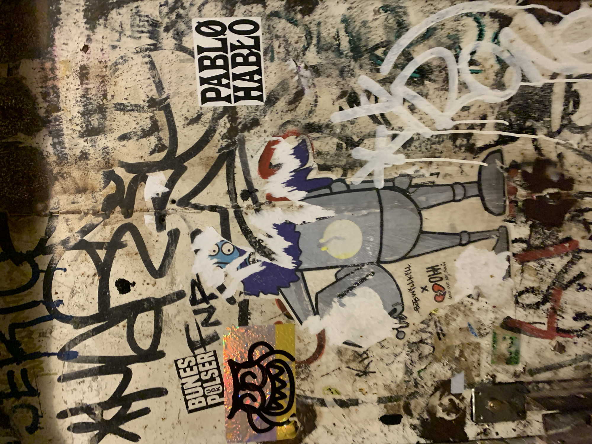 The image shows a section of a graffiti-covered wall in Barcelona, Spain. The wall is primarily light beige with various black and white graffiti markings, stickers, and residue. A sticker with the words "PABLO HABLO" in black block letters is visible. There are several stickers featuring cartoon-like characters. One sticker appears to depict a partially torn figure with blue and red elements. Another sticker shows a metallic figure. Further down, there is a shiny, iridescent sticker with a black design. A sticker with the text "BUNES PULSER" is also visible. Parts of other stickers, faded and torn, are scattered around. The wall shows signs of wear and tear, with scratches and stains.