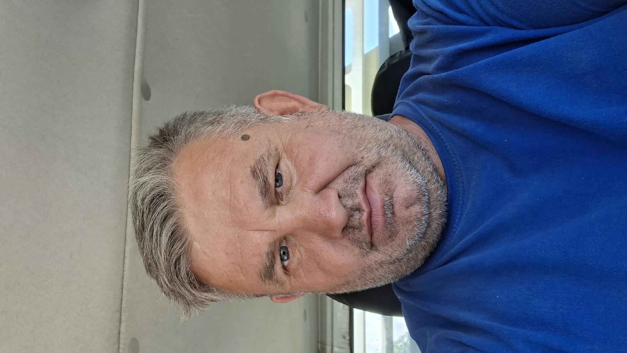 The image shows a close-up, head-and-shoulders selfie of a middle-aged man with graying hair and a beard. He is wearing a blue t-shirt. The man has light blue eyes and a mole on his forehead above his left eye. His expression is neutral. The background appears to be the interior of a vehicle, with part of a headrest visible behind him and what looks like a window to the right. The ceiling of the vehicle is also visible at the top of the frame. The lighting suggests it is daytime, likely outdoors or in a well-lit indoor environment. There is no discernible indication of the specific location within Hîncești, Moldova.