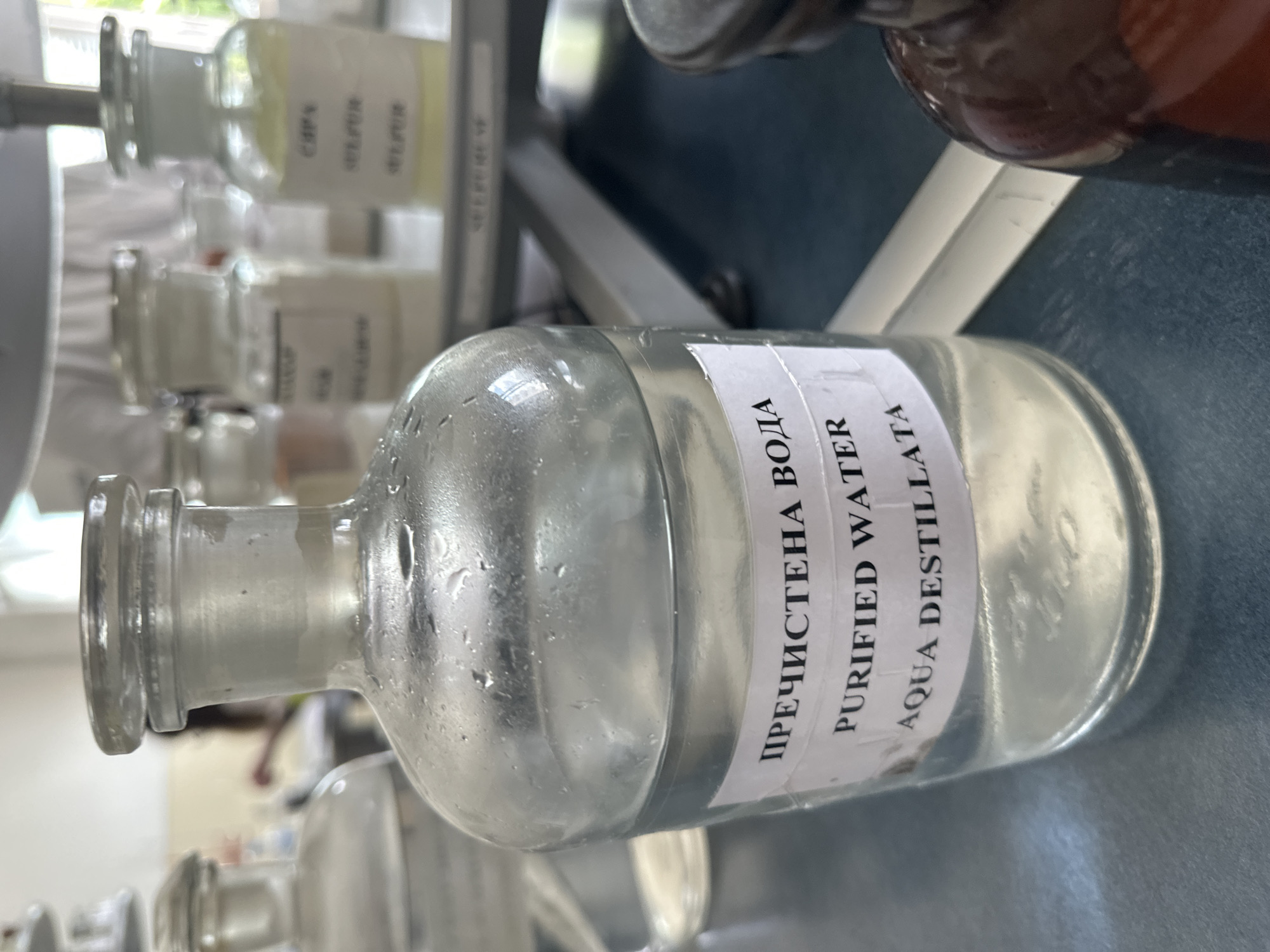 The image captures a clear glass bottle, partially filled with a transparent liquid, resting on a flat, dark surface, likely a laboratory bench or shelf. The bottle's surface is covered in visible condensation or small water droplets.
A prominent white label wraps around the main bottle, detailing its contents in three languages. The top line reads "ПРЕЧИСТЕНА ВОДА," which is Bulgarian for Purified Water. Below this, "PURIFIED WATER" is printed in English, followed by "AQUA DESTILLATA" in Latin. The presence of the Bulgarian text firmly places the scene within Bulgaria, consistent with the provided location context of Sofia.
In the blurred background, several other glass bottles are visible, suggesting a larger collection or storage area. One bottle in the upper left background has a partially legible label with Cyrillic text, appearing to be "СЕРЕБР" (likely part of 'silver' in Bulgarian) and "SILVER" in English, with "COPPER" also faintly discernible below it. Another brown glass bottle is partially visible in the top right corner.
The setting appears to be an indoor scientific or pharmaceutical environment, possibly a laboratory or pharmacy, characterized by the methodical storage of various reagents or solutions. Natural light enters from a window in the upper left background, indicating it is daytime. No people are present, and no specific activity is taking place; the scene depicts items in storage.
