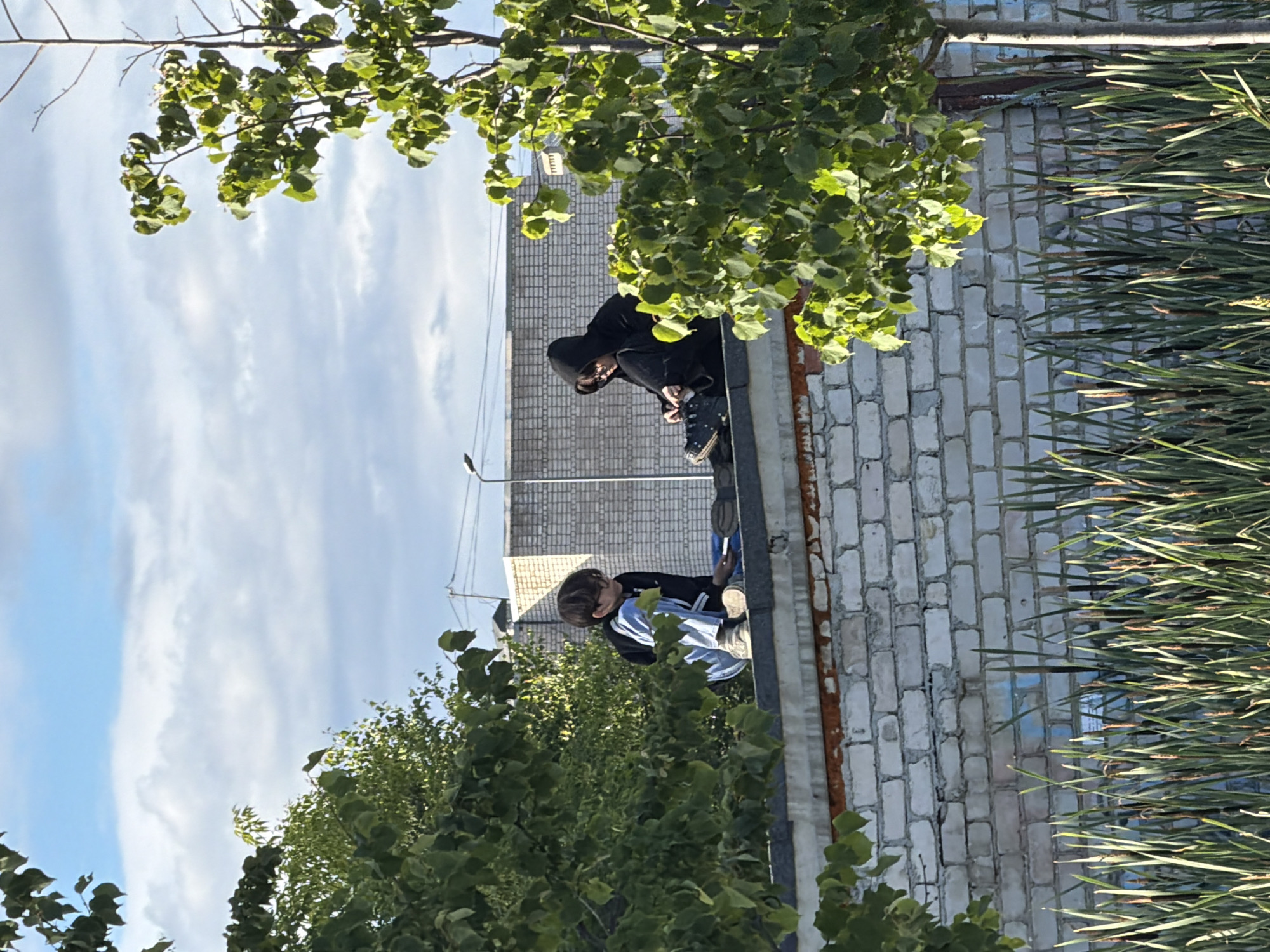 The image shows two people in a semi-urban outdoor setting, likely during the daytime under a clear sky with scattered clouds. The perspective is slightly angled from above, looking down at the subjects.
The first person, closer to the foreground and slightly to the left, is wearing a black hoodie with the hood up, obscuring most of their face. They are crouched down, leaning forward. Visible details include dark pants and what appear to be chunky, dark-colored shoes, possibly platform sneakers or boots, with laces. Their posture suggests they are either examining something on the ground or adjusting their footwear.
The second person, further back and to the right, is also wearing dark clothing, including a jacket or hoodie, and lighter-colored pants, possibly light blue or white. They are also in a crouched or seated position, facing away from the viewer. A blue and white garment is visible around their torso, and they appear to be wearing white sneakers.
Both individuals are positioned on what looks like the edge of a building or elevated structure, possibly a rooftop, with a white brick wall extending to the right. There is a rusty metal element running along the edge of the structure. Lush green foliage, including tree branches and leaves in the foreground and midground, partially obscures the view of the subjects and the background. Some tall, thin, green plants are also visible on the right side of the image.
The scene appears casual and perhaps adventurous, with the subjects in a somewhat unconventional and elevated location. The lighting suggests it's daytime, likely morning or afternoon. No text is visible in the image.