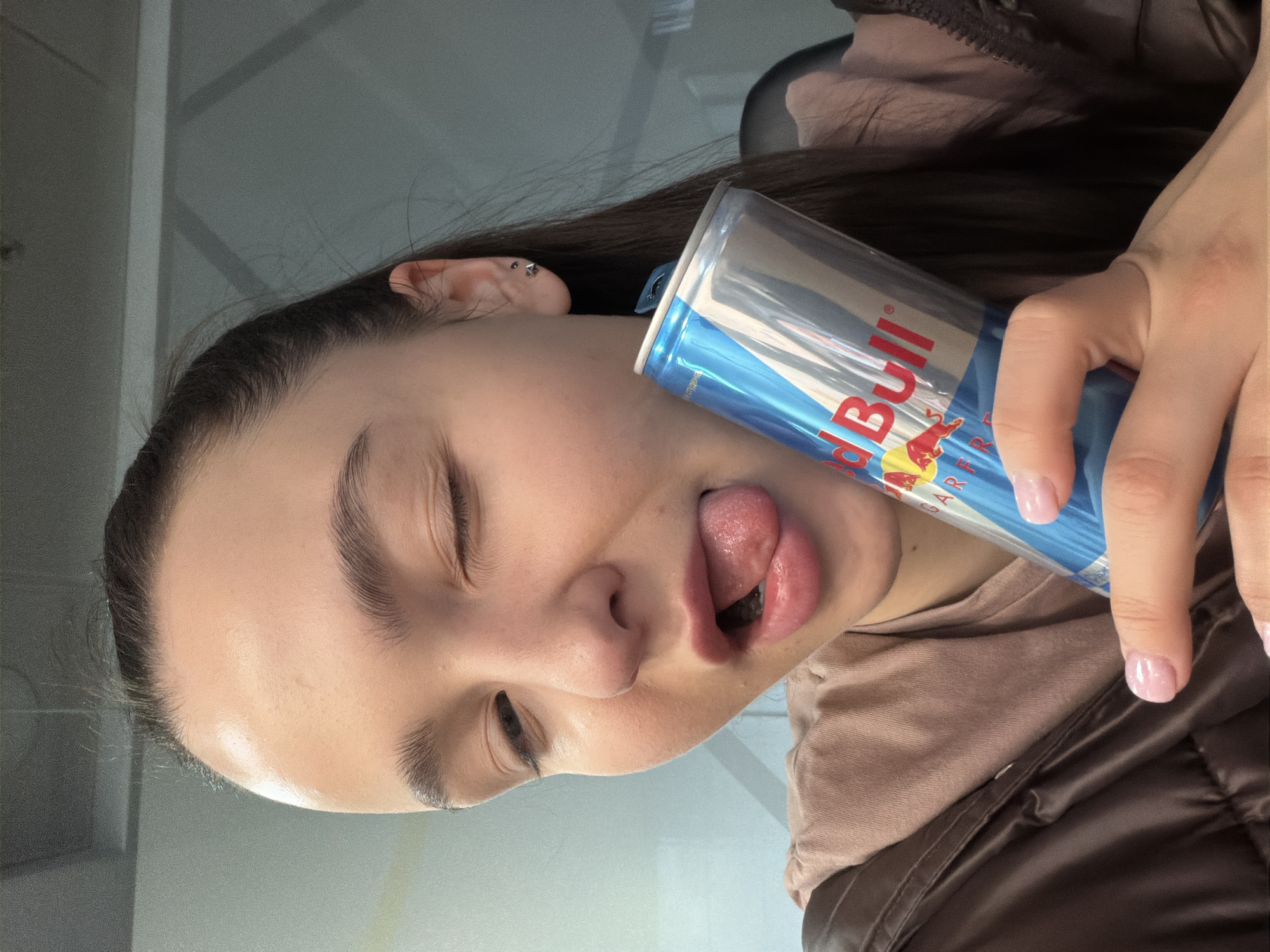 A young adult woman is positioned sideways in the frame, holding a blue and silver Red Bull energy drink can against her face. She has dark hair and fair skin. Her eyes are closed and her tongue is sticking out. She is wearing a tan shirt and a brown jacket. The background is a neutral gray, suggesting an indoor environment. No specific information indicates this image was taken in Sofia, Bulgaria.
