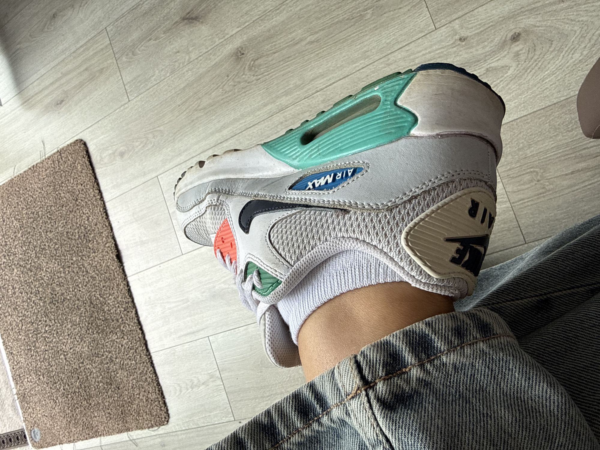 The image displays a close-up view of a person's lower legs and feet, with a focus on a Nike Air Max sneaker. The sneaker is predominantly white with accents of red, green, and turquoise. The person is wearing distressed denim jeans and white socks. The footwear is positioned on a light-colored wooden floor, partially overlapping a textured, brown doormat. The scene appears to be indoors. The city of Heiligenrode, Germany, is not discernible from the visual elements present in the image.