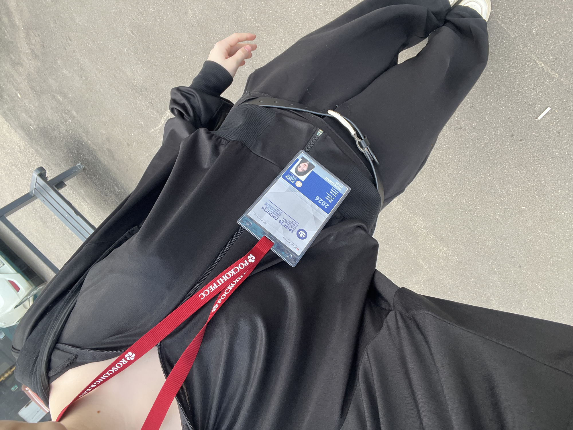 A first-person, high-angle shot captures a person looking down at their torso and legs, likely outdoors on a paved surface. The individual is dressed in an all-black athletic or casual outfit, consisting of a jacket, pants, and white sneakers. A black belt with a silver buckle is worn around the waist.
Prominently displayed is an identification badge, hanging from a red lanyard that features the "РОСКОНГРЕСС" (Roscongress) logo and "ROSCONGRES" branding. The badge identifies the event as "SPIEF '26" or the "ST. PETERSBURG INTERNATIONAL ECONOMIC FORUM" (ПМЭФ 26), scheduled for "2026." The name "Мария Guseva" (Mariya Guseva) is visible, along with her role as "STAFF" (ПЕРСОНАЛ). Other text on the badge includes "FORUM-SPERU."
The setting appears to be a daytime outdoor environment, with gray asphalt or concrete underfoot. A white cigarette butt is discernible on the ground near the person's feet, and a metallic structure, possibly part of a bench or railing, is visible in the lower left background. The visible text on the badge and lanyard strongly suggests the person is attending or working at the St. Petersburg International Economic Forum in Russia.